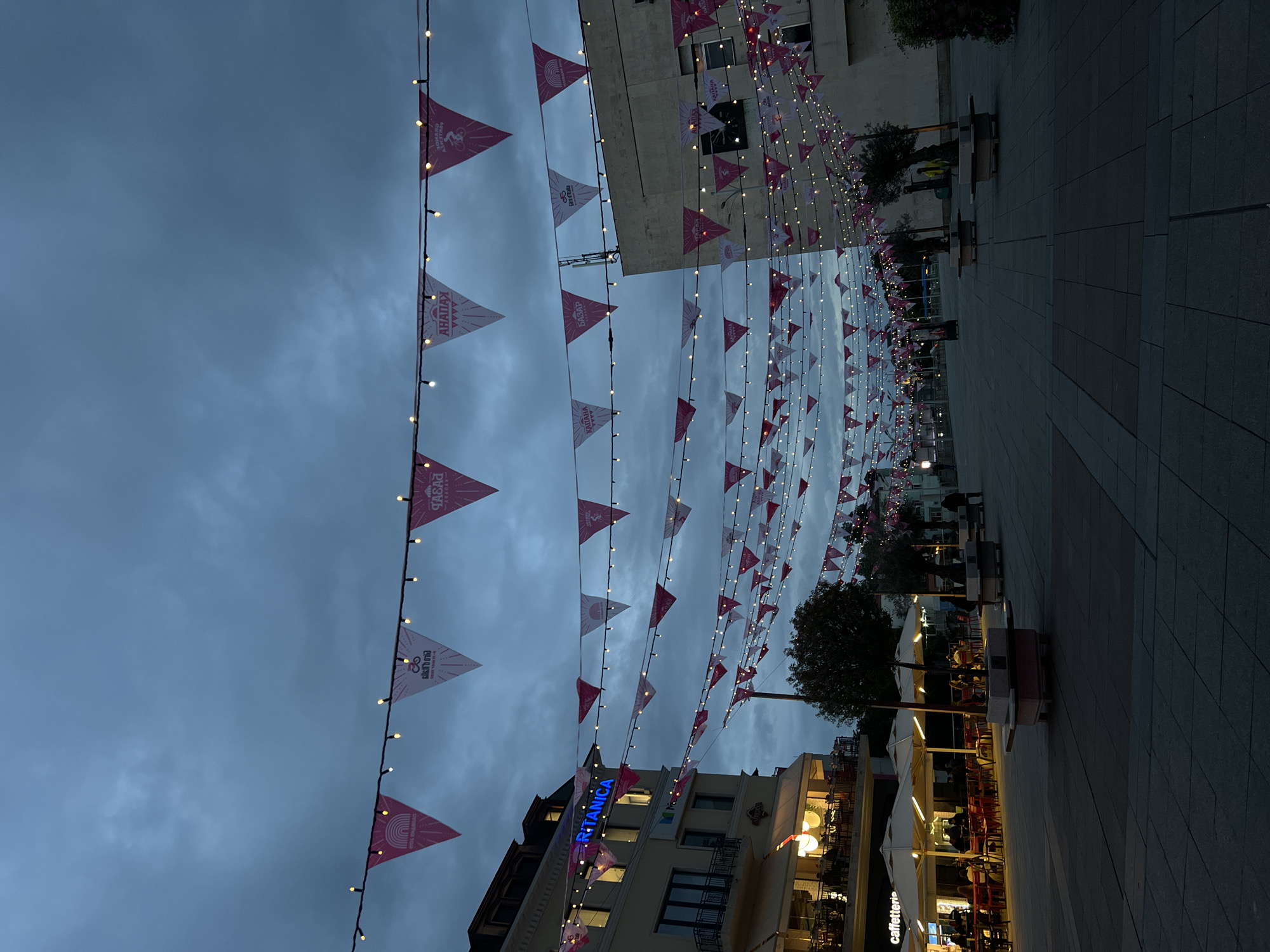 The image captures a pedestrianized street scene in Plovdiv, Bulgaria, at dusk. Rows of triangular flags with light bulbs strung along the top are suspended above the street, creating a festive atmosphere. The flags display a recurring design with pink and white colors. The street is paved with light gray tiles. On the right, there are benches and trees lining the pedestrian path. Further down the street, outdoor seating areas of cafes and restaurants are visible, illuminated by warm light. On the left, buildings stand, including one with a blue "BRITANICA" sign. The sky is overcast with dark, heavy clouds. People are present in the scene, primarily seated at the outdoor cafes and restaurants.