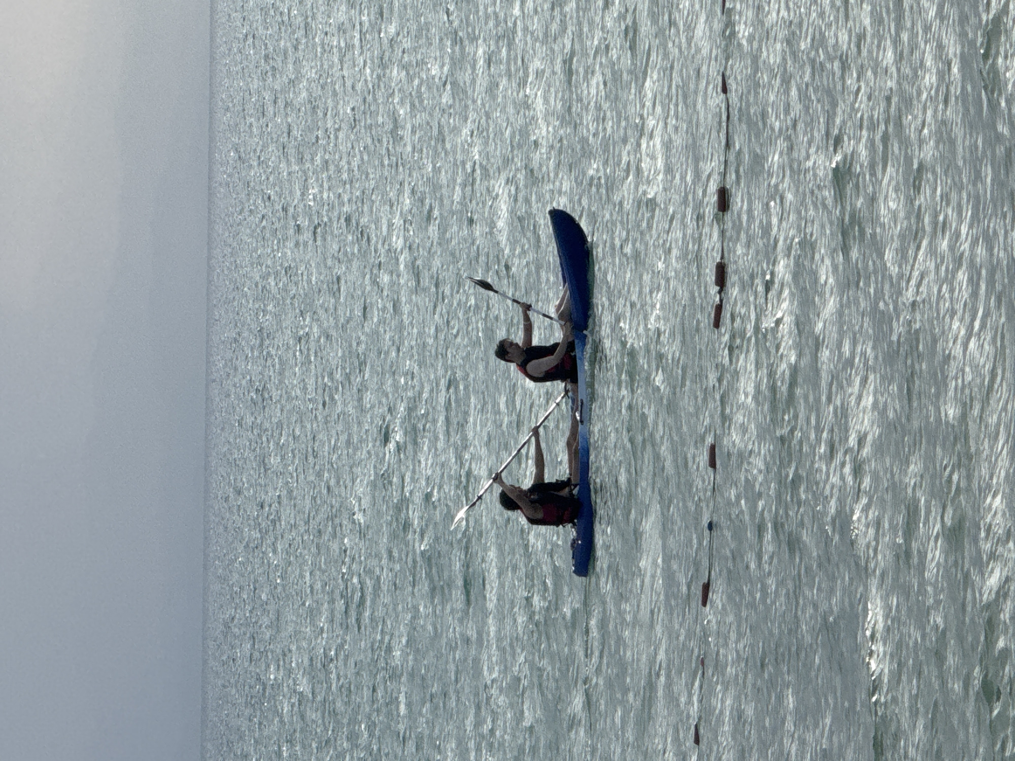 Two people are kayaking on a body of water under an overcast sky. The kayaks are tandem, with one person in front and the other behind. Both individuals are wearing life jackets and holding paddles. The water is choppy, with small waves and sunlight glinting off the surface. In the foreground, there is a series of buoys floating on the water. The background is a hazy, gray sky, suggesting it may be a cloudy day or dawn/dusk. The scene depicts a recreational activity on a large body of water, possibly a lake or the sea. The specific location within Sary-Oy, Kyrgyzstan, cannot be determined from the visual information alone.
