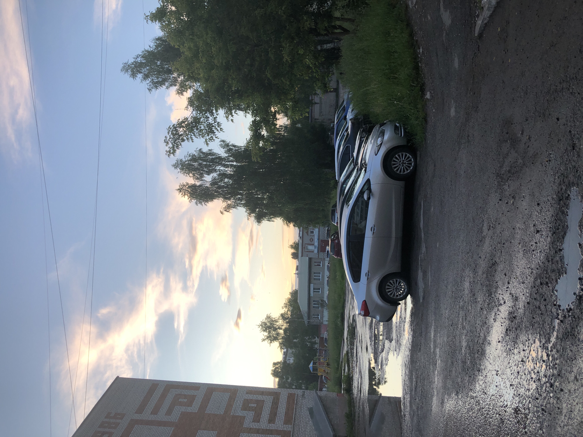 The image depicts a parking lot with several cars parked in front of a building and a grassy area. The sky above is partly cloudy with a sunset glow. In the foreground, a silver sedan is parked on a wet, gravelly surface with several puddles. To the left, a portion of a brick building with geometric patterns is visible. In the background, there are more cars, trees, and a low-rise building. A playground structure can be seen in the distance behind some trees. The scene appears to be a residential or municipal area. The photograph was taken in the city of Solikamsk, Russia.