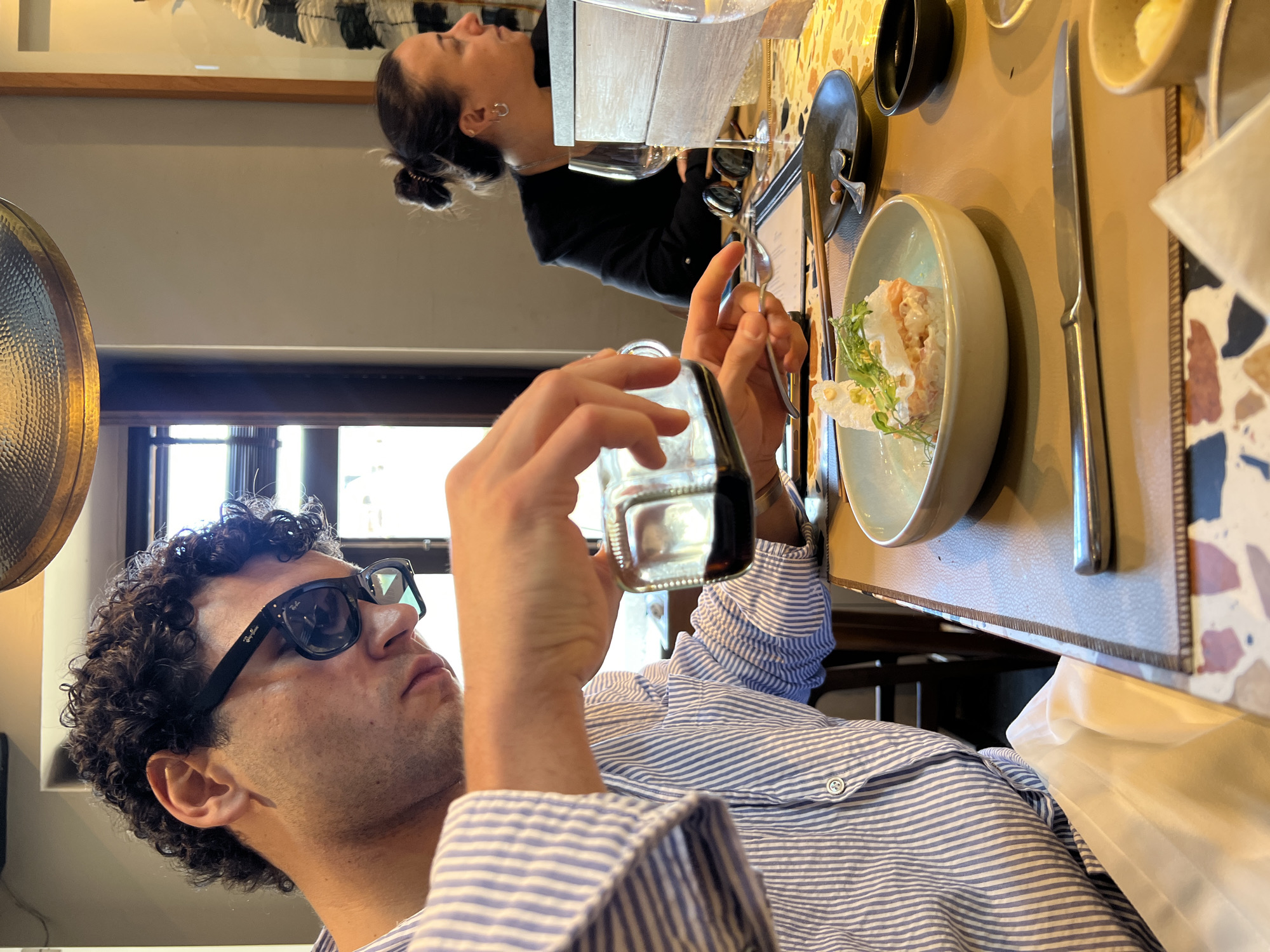 A man with curly hair and sunglasses is taking a photograph of a plate of food with a small glass jar in a restaurant. He is wearing a light blue and white striped shirt. The plate of food contains a pale pink mixture with green herbs and white crisps. The table is set with a black bowl, a knife, and a menu. The table has a beige placemat. A woman with her hair up in a bun is in the background. The restaurant appears to be indoors with natural light coming in through a window. The exact location in Porto, Portugal is not apparent from the image.