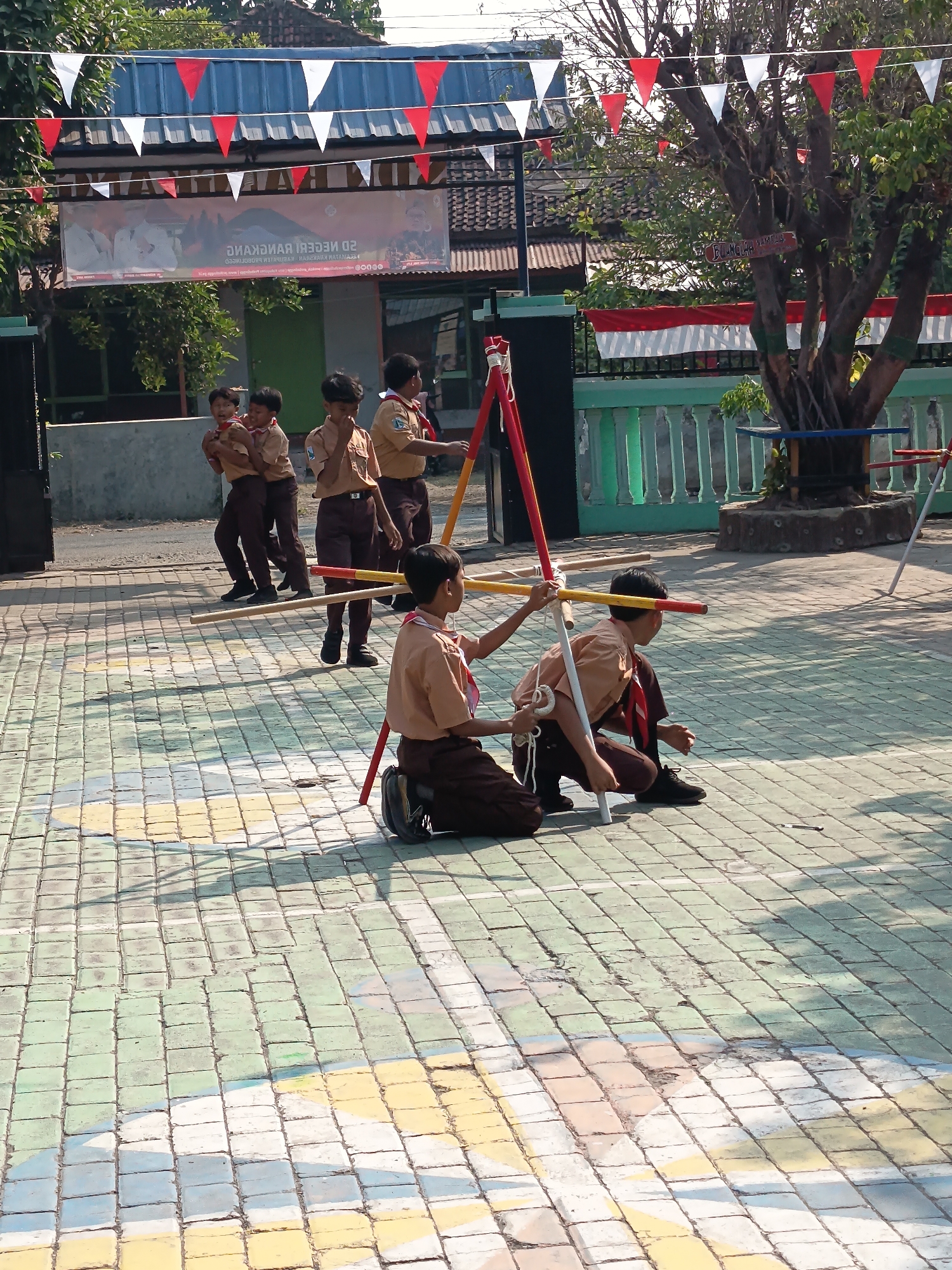 In the city of Gebangan, Indonesia, a group of young students in scout uniforms are engaged in an outdoor activity on a paved ground. The pavement is decorated with colorful square tiles in green, blue, yellow, and white. The students, dressed in khaki shirts and brown pants with red neckerchiefs, appear to be participating in a game or a lesson involving a wooden structure with red and yellow poles.

In the foreground, two boys are in a kneeling or squatting position, interacting with the wooden structure. One boy, on the right, is facing away from the camera, looking towards the ground. The other boy, on the left, is also in a similar pose, his hands near the wooden poles.

Further back, a group of four boys in scout uniforms are standing and walking. They are also wearing khaki shirts and brown pants with red neckerchiefs. One boy in the back right is partially obscured by the wooden structure.

The background shows a building with a blue roof, adorned with red and white triangular flags strung across the front. A banner with Indonesian text is also visible on the building. To the right, a large tree with visible roots stands next to a green fence. More red and white flags are displayed on the fence and on the tree. The overall atmosphere suggests a school event or an outdoor educational activity.