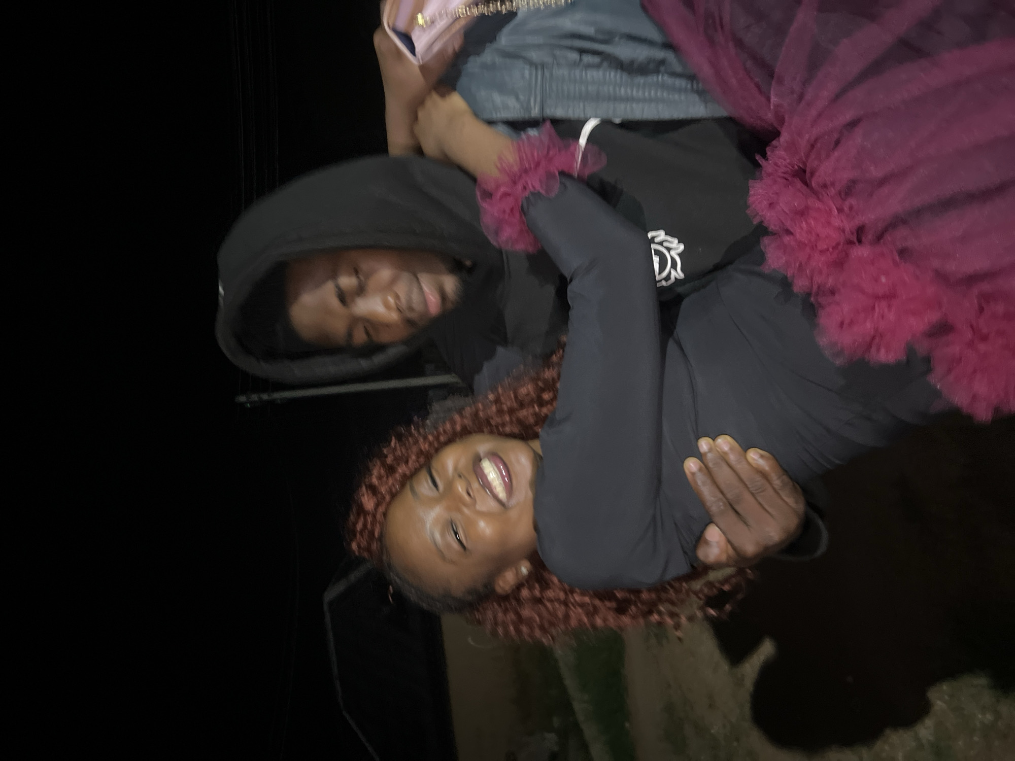 A young Black man in a black hoodie carries a young Black woman in his arms, who is wearing a black dress and a magenta tutu-like skirt. Both are smiling broadly. The man appears to be in his late teens or early twenties, and the woman is likely of a similar age. They are outdoors at night, with a dimly lit building and a utility pole visible in the background. The setting is likely a residential area in Durumi, Nigeria, given the style of the building and the overall ambiance. The image captures a moment of shared joy and affection between the two individuals.