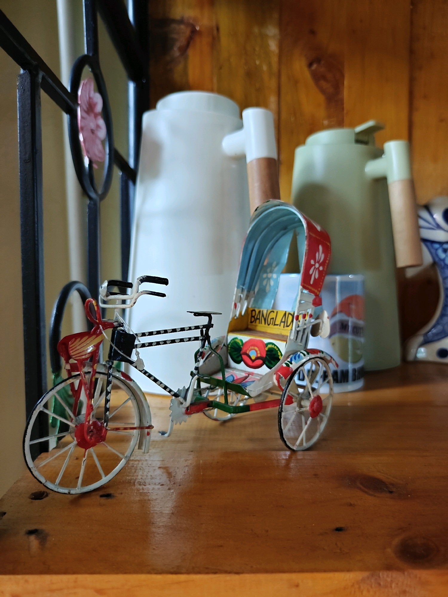 A miniature, colorful rickshaw toy is displayed on a wooden shelf. The rickshaw is detailed with red, white, green, and blue accents, featuring a canopy and wheels. The word "BANGLAD" is visible on the side of the rickshaw, suggesting it might be a representation of a Bangladeshi mode of transport. In the background, two large, light-colored pitchers and a decorative blue and white ceramic piece are visible. The shelf is made of natural wood with visible grain. The scene appears to be an indoor display. The location is not specified in the image.