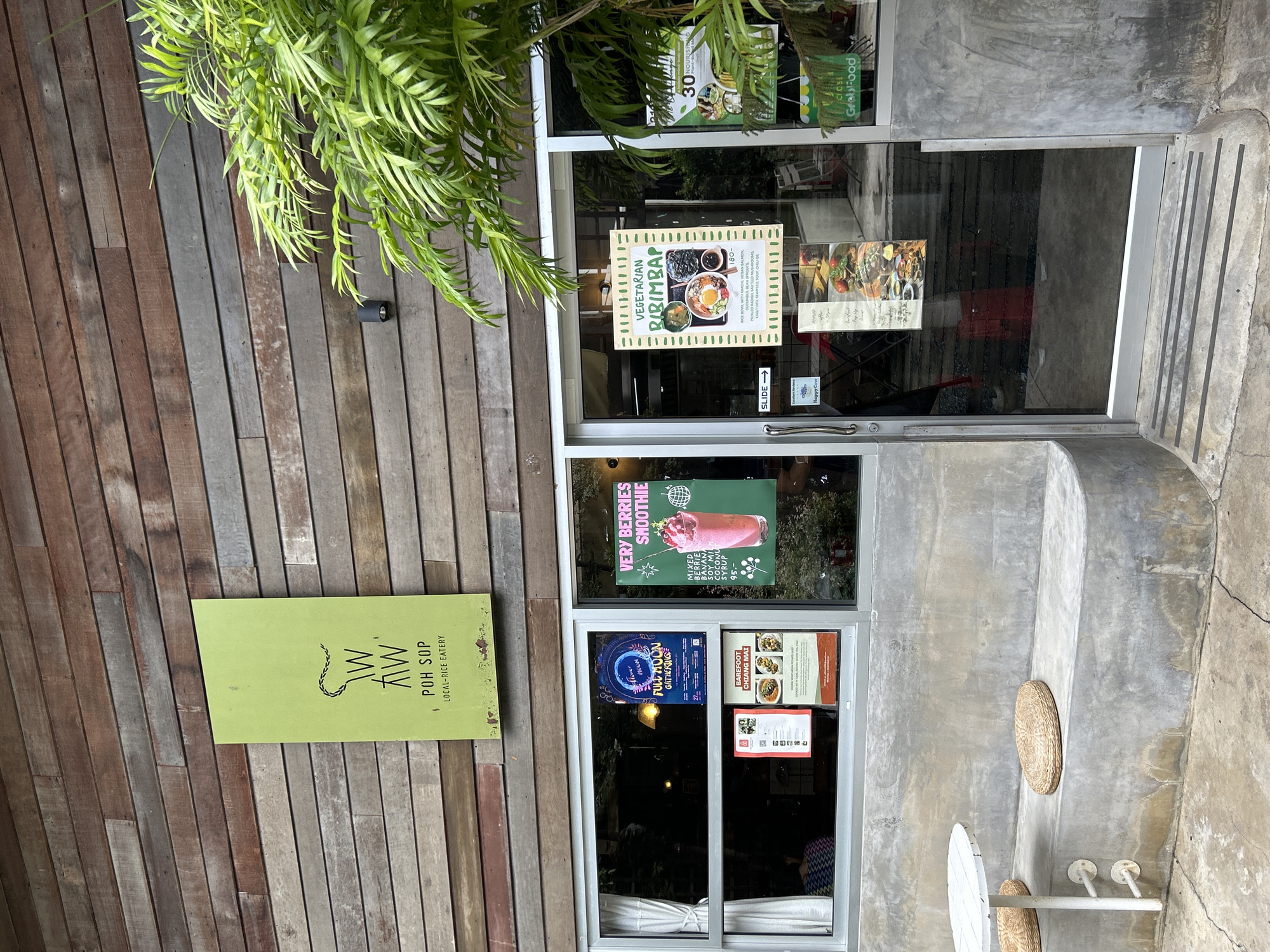 The image shows the exterior of a restaurant named "AW Poh Sop Local-Rice Eatery" in Chiang Mai, Thailand. The building has a wooden facade with horizontal planks. A lime-green sign with the restaurant's logo and name is mounted on the upper part of the building. Below the sign, a large glass storefront displays various posters advertising menu items such as "Very Berries Smoothie" and "Vegetarian Bibimbap." There is also a poster for "Full Moon Gathering." Inside the restaurant, reflections in the glass show tables and chairs. To the left of the entrance, a concrete bench with two woven cushions and a white table are visible. To the right, lush green foliage from a palm tree partially obscures the storefront. A sign on the door reads "SLIDE." The overall environment appears to be a bright, possibly daytime scene.