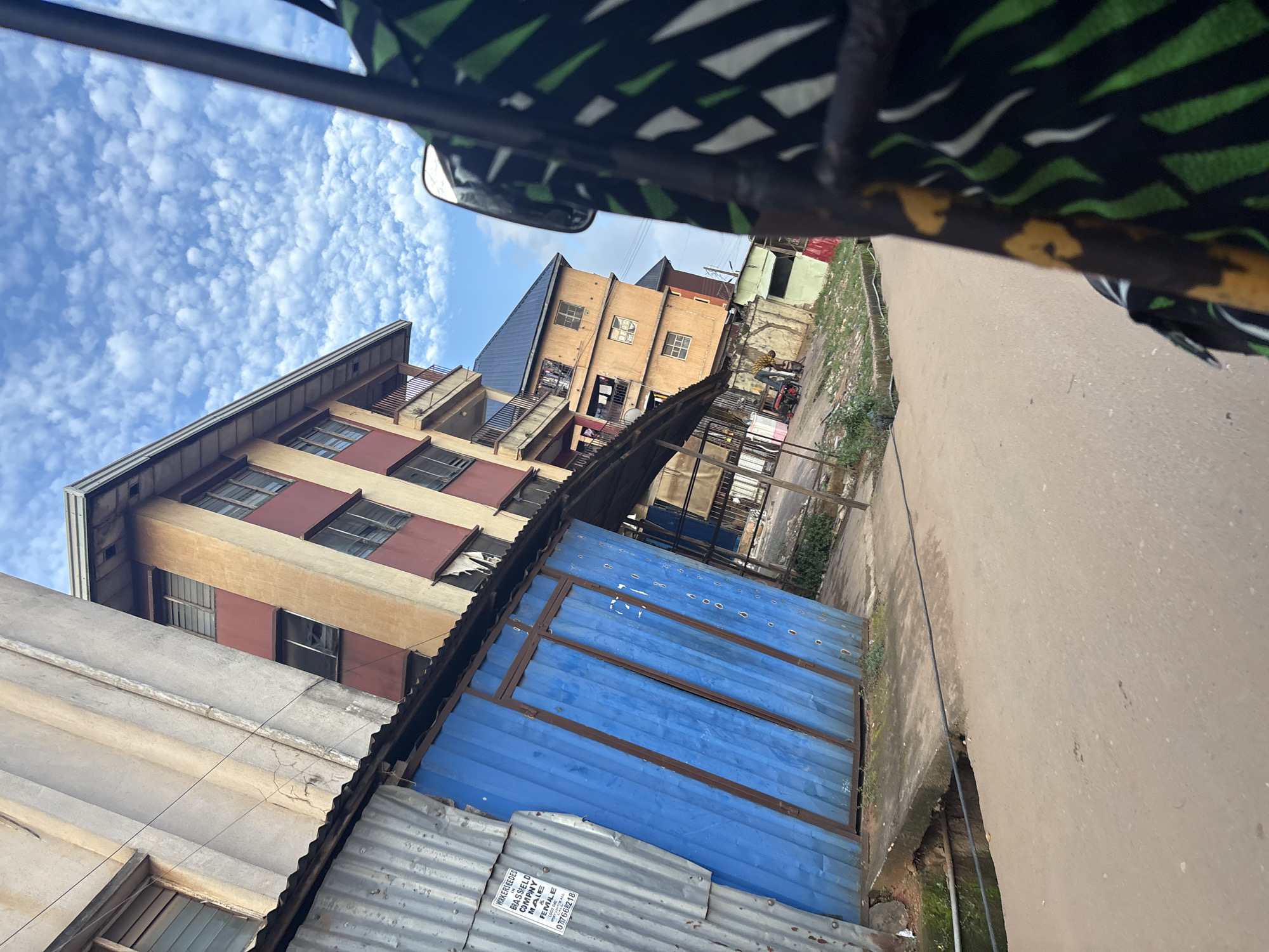 The image depicts a street scene in Onitsha, Nigeria, viewed from the perspective of someone inside a vehicle. In the foreground, the right side of the frame is dominated by the patterned upholstery of the vehicle's interior, featuring a black base with repeating green, white, and yellow abstract shapes. A dark metal bar, likely part of the vehicle's structure, runs vertically through the foreground.
The background reveals a mix of urban structures. To the left, a corrugated metal structure with a blue corrugated metal door is prominently featured. A sticker with text, including "MAKERSSEED BIASFIELD COMPANY MALE" and a phone number, is affixed to the corrugated metal wall of a adjacent structure. Beyond this, a multi-story building with reddish-brown and pale yellow facades stands at an angle, its windows visible. Further down the street, another, more modern, building with a dark roof and yellow and reddish-brown sections is visible. The sky above is filled with scattered white clouds against a bright blue backdrop. The street surface is paved with a light brown asphalt material, and there appears to be a drainage channel or ditch on the left side of the street with some debris. In the distance, a few people and what appear to be motorcycles or tricycles are visible, suggesting ongoing activity in the area. The overall impression is of a bustling, everyday street in a Nigerian city.