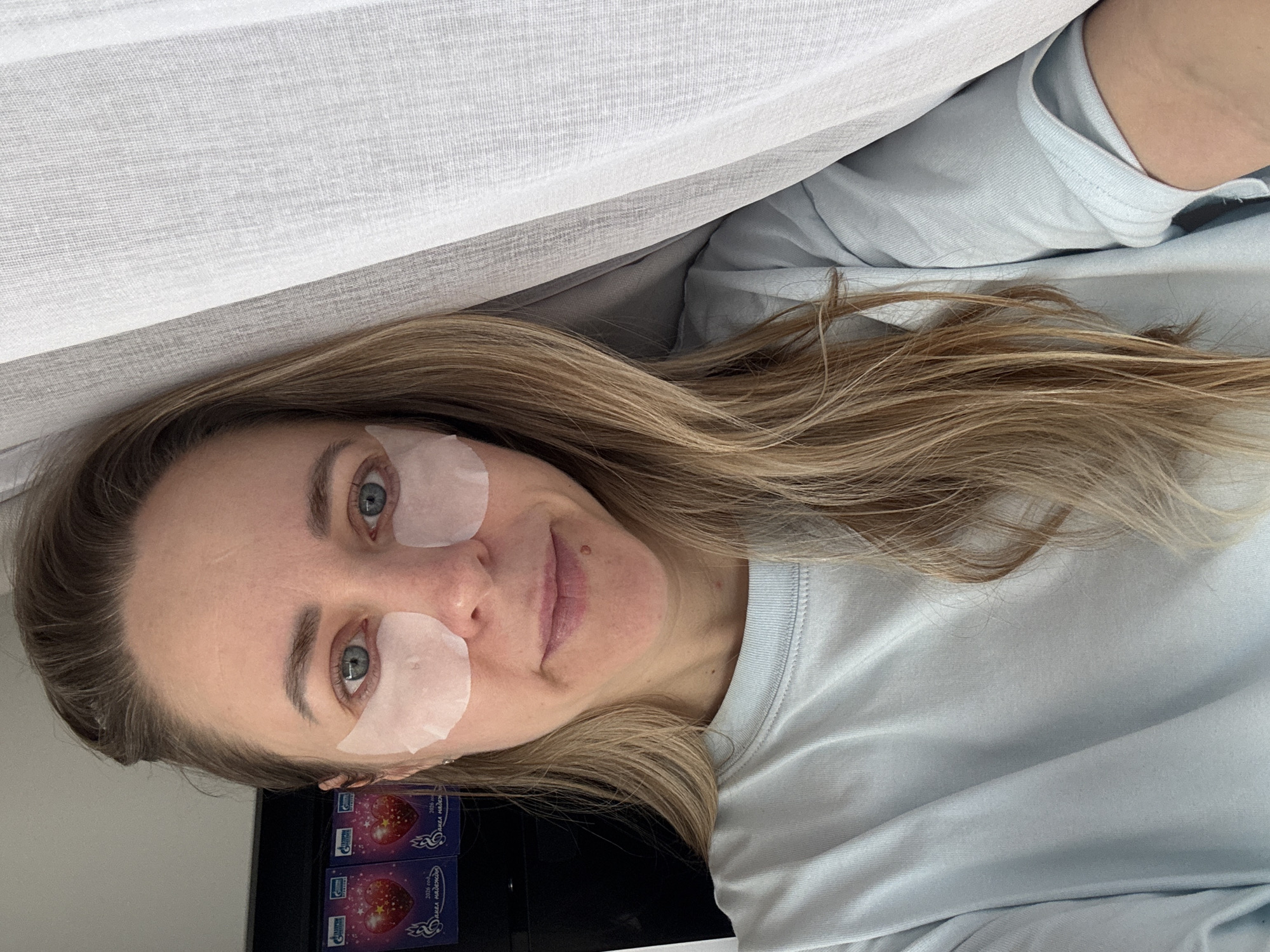 A fair-skinned woman with light brown hair is lying on a light-colored surface wearing white under-eye patches. She is wearing a light blue shirt. A white curtain or fabric is draped above her. Two small blue and red boxes are visible in the lower left corner of the image. The location is suggested to be Chaikovsky, Russia based on the text on the box.