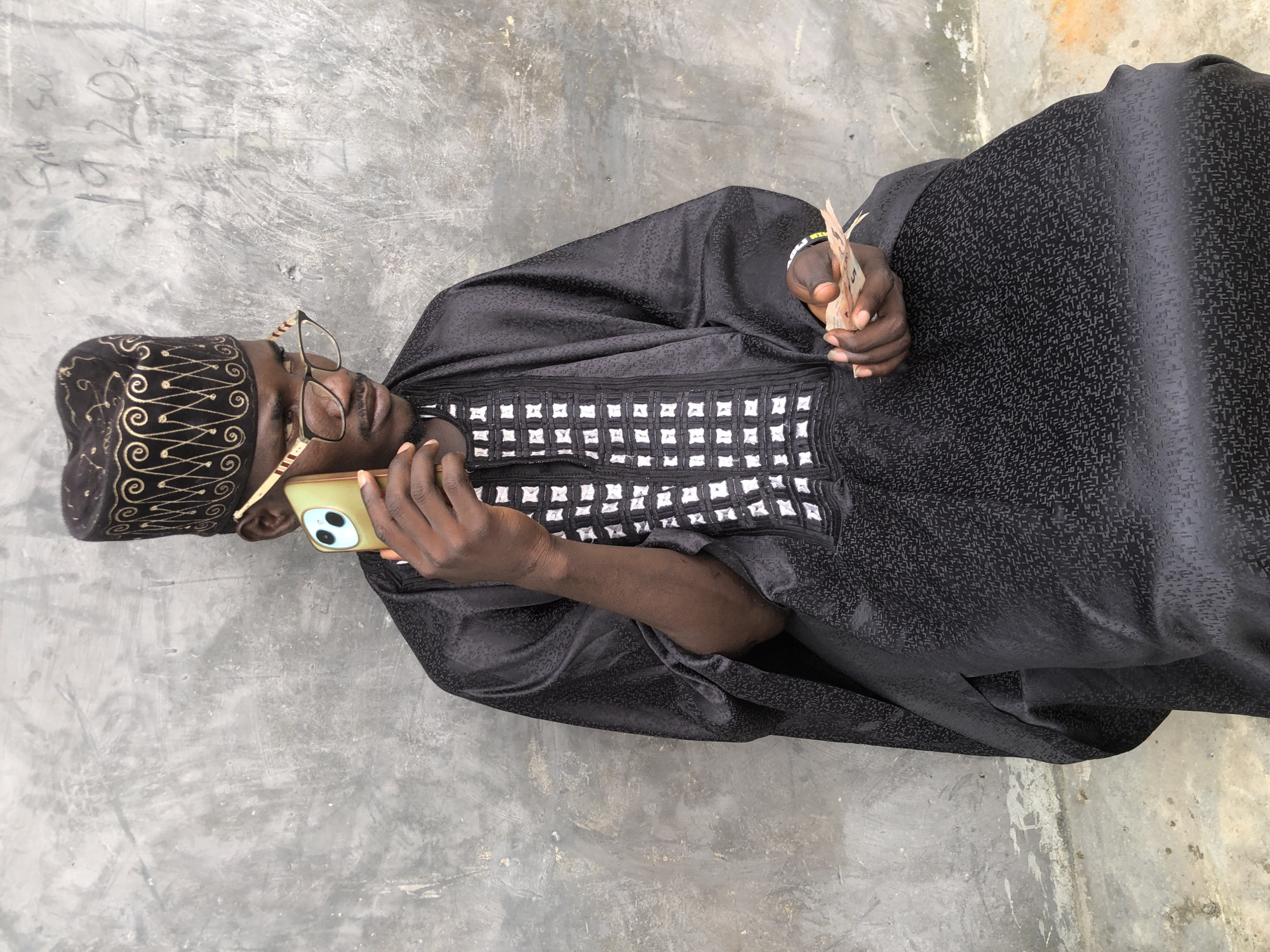A young Black man wearing a black traditional cap and black robe is sitting in front of a textured gray wall. He is holding a gold-colored smartphone to his right ear with his right hand. His left hand is holding a small wad of folded banknotes. The man is wearing eyeglasses with gold-colored frames. The robe has white patterned embroidery on the chest. The background wall has some faint graffiti or markings on it, including what appears to be "1920s". The ground beneath him appears to be concrete, with some scattered debris. The overall setting suggests an outdoor urban environment.