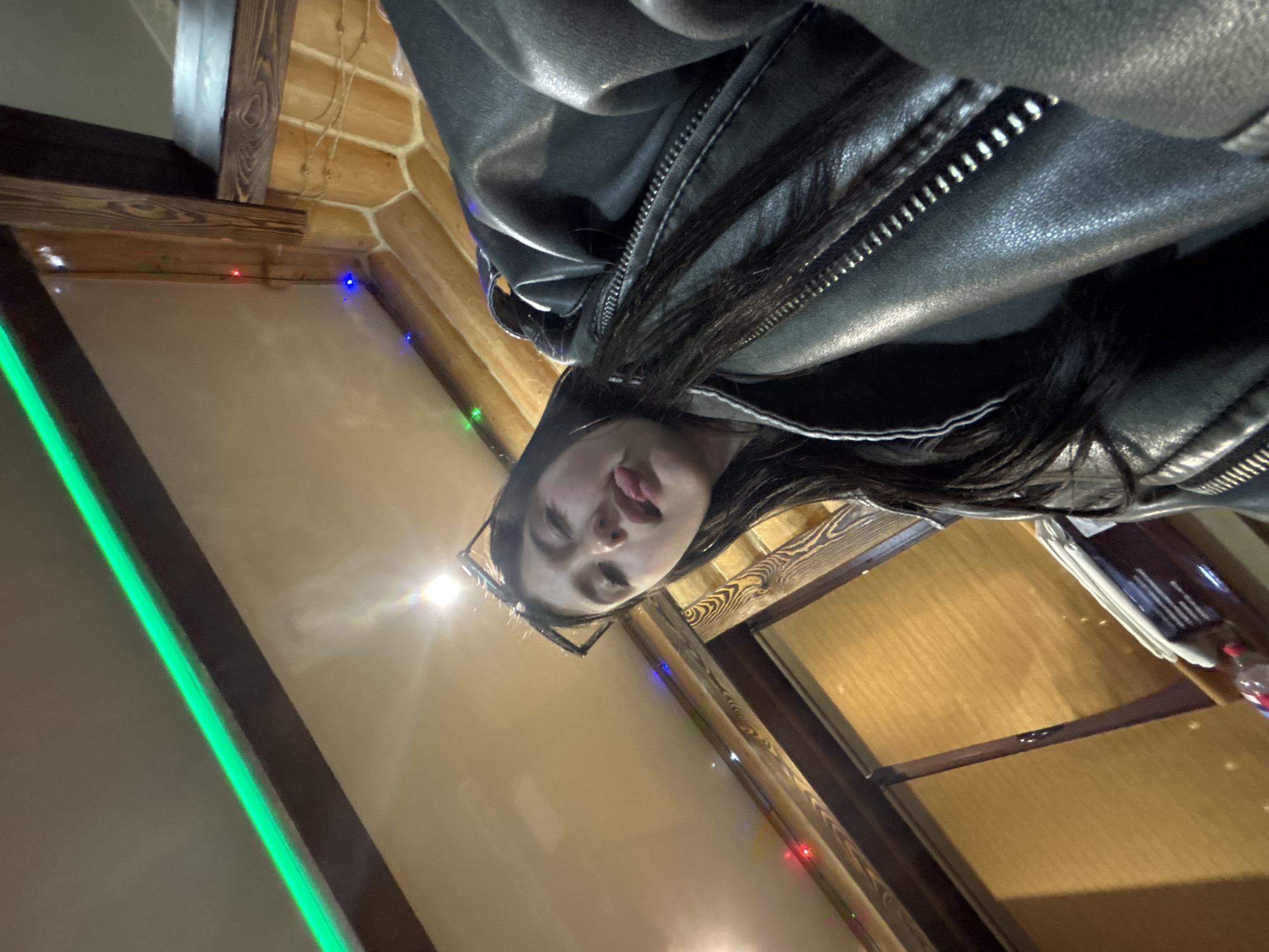 A woman with long dark hair and glasses is seen from a low angle, wearing a black leather jacket. She has her tongue slightly out. The background includes wooden walls or paneling, with festive colored lights strung along the edges. A green light strip is visible on the left side. There is a table with items on it in the background, possibly including a menu and a bottle. The location is likely a restaurant or bar in the city of Chaikovsky, Russia, based on context clues.
