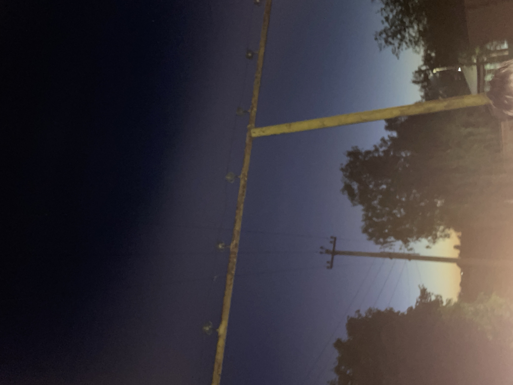 The image depicts an outdoor scene at dusk, with a deep blue sky transitioning to lighter hues near the horizon. A wooden beam, likely part of a power line structure, stretches horizontally across the upper portion of the frame. Several insulators with what appear to be small lights or bulbs are attached to this beam. A wooden utility pole with crossbars and wires stands vertically in the mid-ground. Silhouettes of trees are visible on the left and right sides of the frame, with the sky showing a faint orange glow near the horizon, suggesting the last light of sunset. Some distant structures and a faint light are discernible behind the trees. The lower part of the image is blurred and appears to include a person's head and shoulders, indicating the photograph was taken from a low vantage point. The overall atmosphere is serene and dusky.
