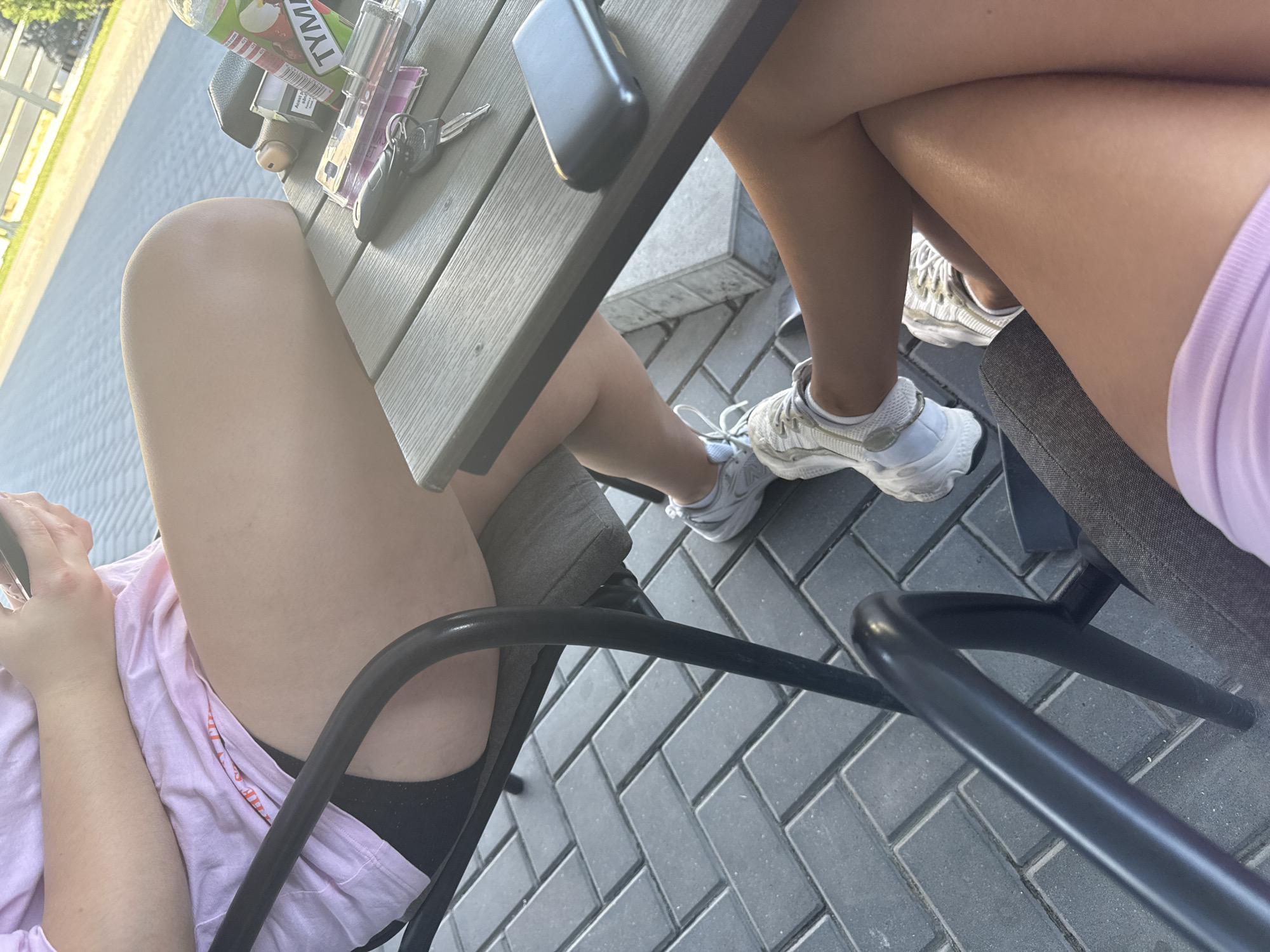 The image shows two individuals, seen from the waist down, sitting at a table outdoors. The person on the left is wearing a pink t-shirt and black shorts, with white sneakers. They are holding a smartphone. The person on the right is wearing white sneakers and a light purple top, with their legs crossed. On the table, there are car keys, a wallet, and a bottle of juice. The table is made of wood and is placed on a patio paved with dark gray rectangular bricks. The background shows a sunlit area with a white fence and some greenery, suggesting an outdoor dining area or park. The scene is likely in Comrat, Moldova, as indicated by the provided context.