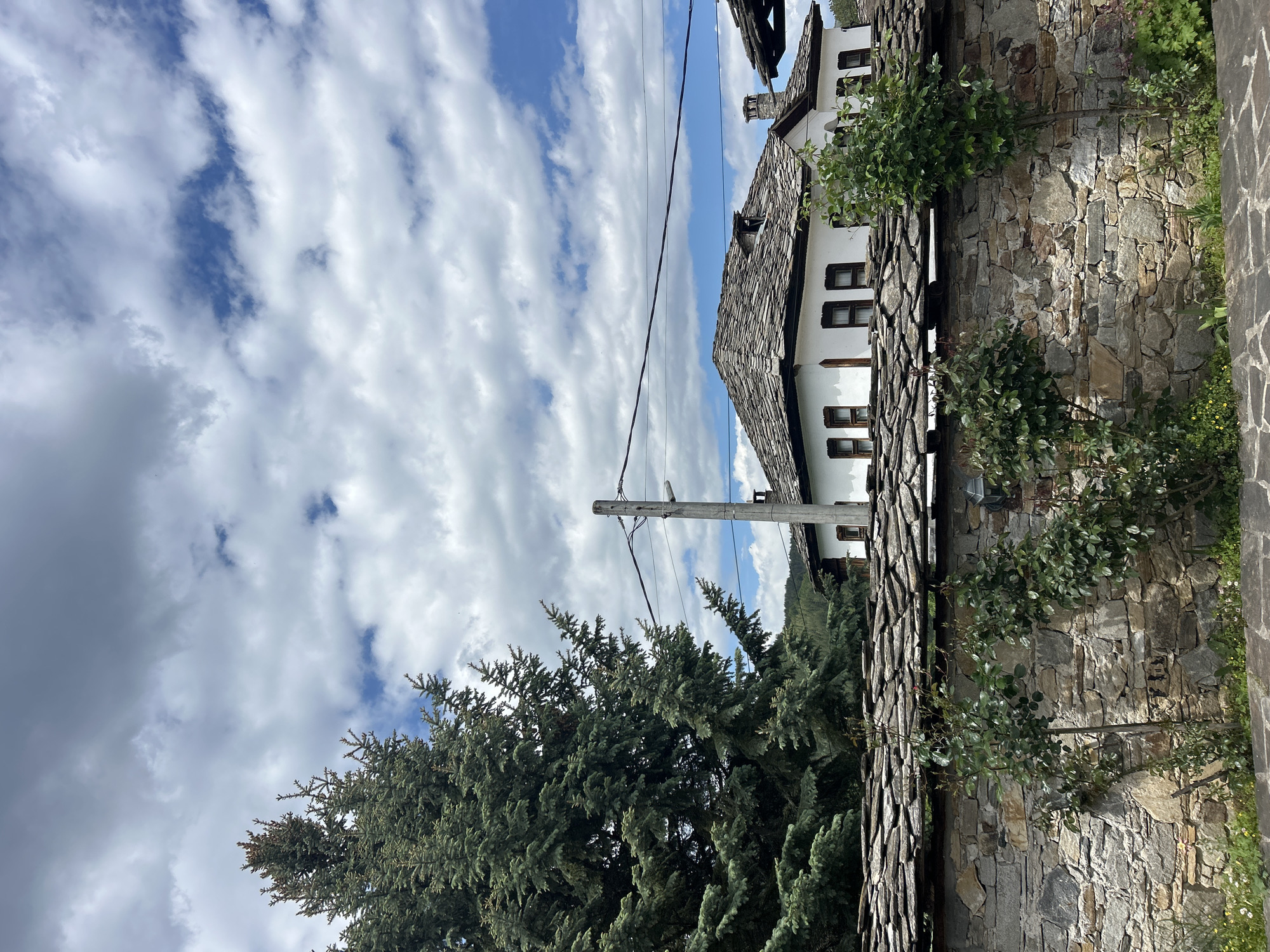 The image depicts a scene in Dolen, Bulgaria. A stone wall with a slate-covered top runs along the right side of the frame. Behind the wall, a white building with dark-framed windows and a slate roof is partially visible. Power lines run from a utility pole in the mid-ground to the building. To the left of the wall and pole, a large evergreen tree dominates the lower portion of the image. The sky is filled with white and gray clouds with patches of blue visible.