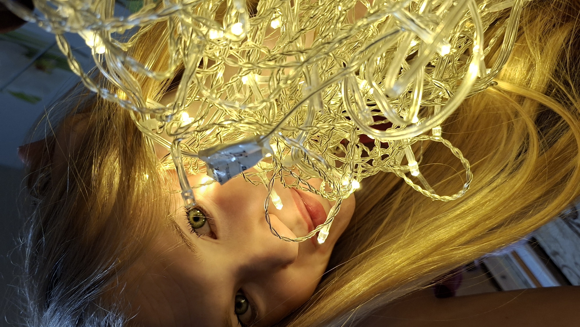A close-up, low-angle shot shows a young woman with long, blonde hair. She is lying down, facing upward. A tangle of lit fairy lights are draped over her face and head, obscuring some of her features. Her left eye is visible, and a portion of her mouth is slightly open. Some of her hair is spread out around her head. The environment seems to be indoors, possibly a bedroom, with hints of furniture visible in the background. There is nothing indicating this image location to be in the city of Большой Букор, Russia.