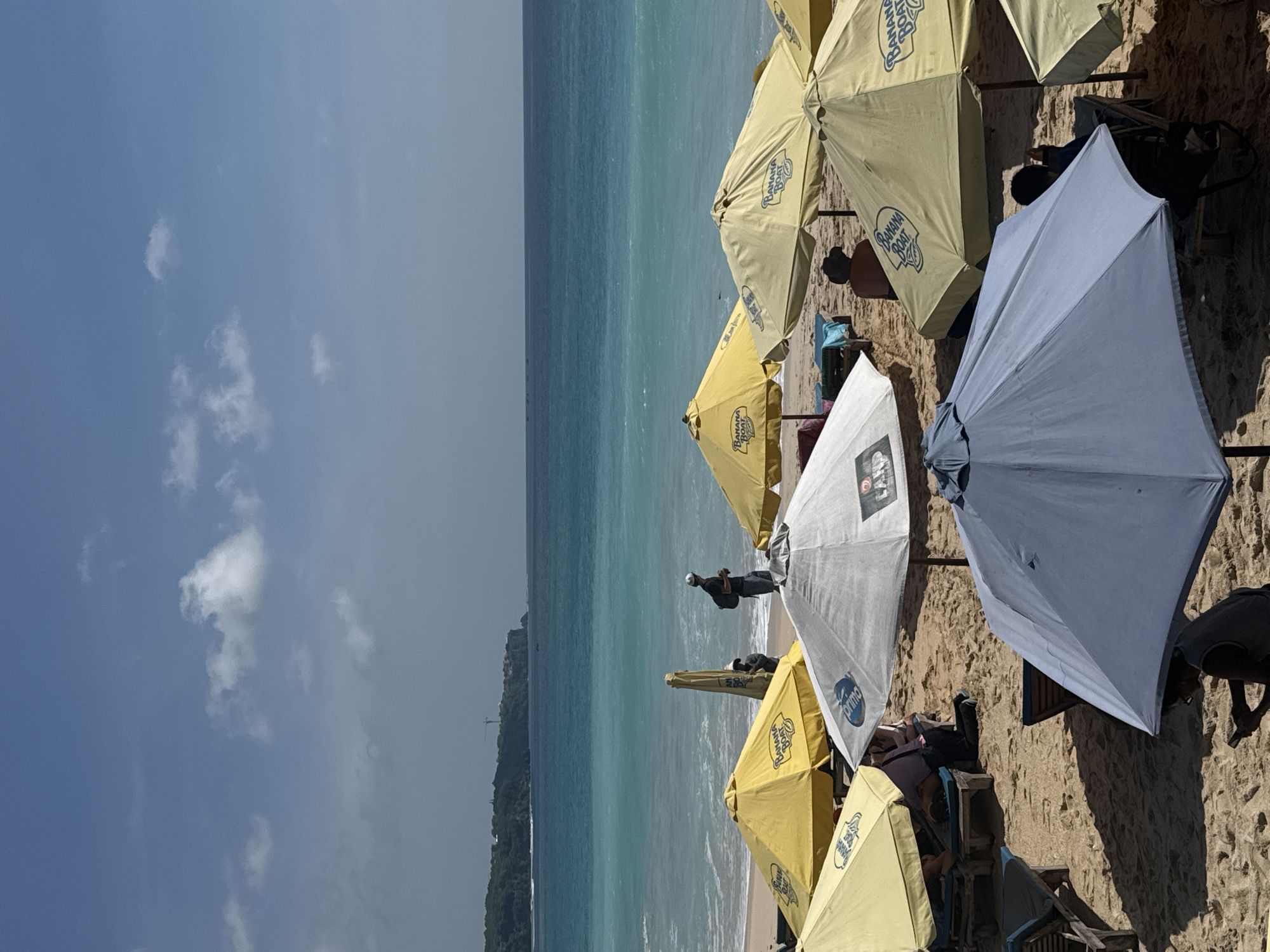 This image captures a beach scene in Kuta Selatan, Indonesia, featuring a row of beach umbrellas shading beachgoers from the sun. The umbrellas are primarily yellow and white, with "Banana Boat" and "Primo" logos printed on them. Beneath the umbrellas, people lounge on chairs. Beyond the umbrellas, the turquoise ocean stretches towards the horizon where a strip of land covered in dense vegetation is seen. The sky is pale blue with scattered white clouds.