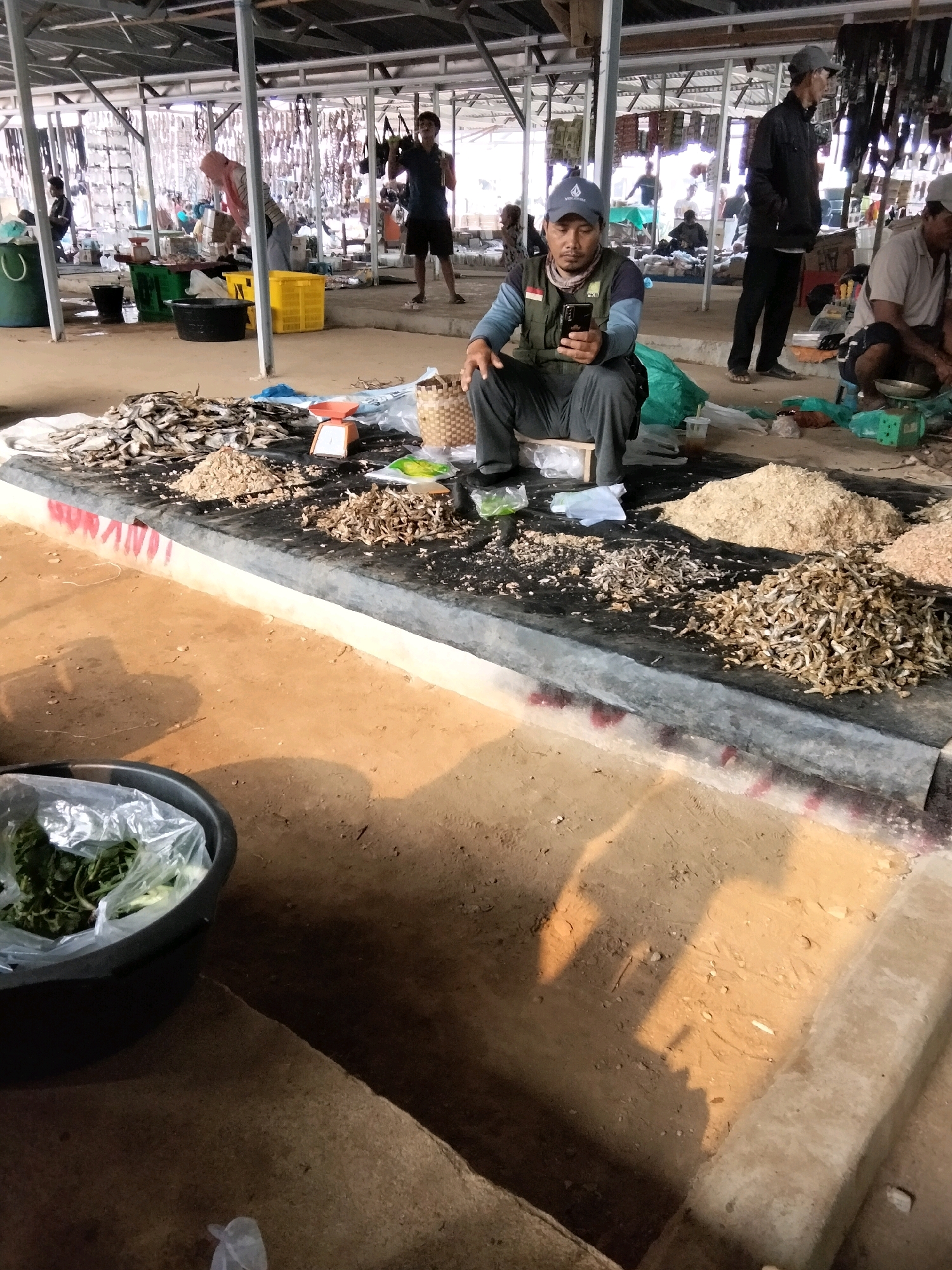 A man wearing a grey baseball cap, a dark vest over a long-sleeved blue shirt, and grey pants is sitting on a wooden stool in front of a display of dried fish at an outdoor market. He is holding a black smartphone in his right hand. The market appears to be located in Tirto Sari, Indonesia. The ground is dirt, and the market is covered by a corrugated metal roof supported by metal poles. In the background, other people are visible, as well as various market stalls and goods, including what appear to be more dried fish, baskets, and crates. A large black tub containing green vegetables in plastic wrap is in the foreground. Sunlight casts shadows across the ground.