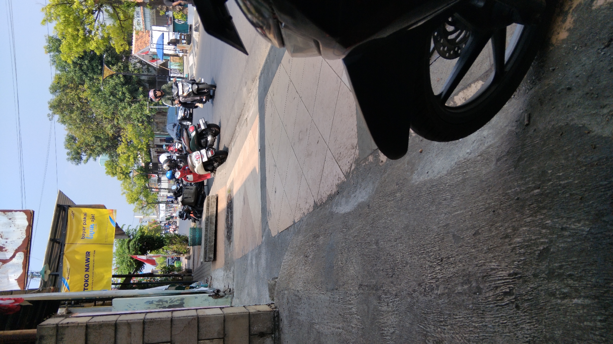 The image depicts a street scene in Batang, Indonesia, likely in the vicinity of Toko Nawir, as indicated by a prominent yellow banner. The banner advertises "Toko Nawir" and "bayar pakai livin aja!", suggesting a retail store that accepts payments via the Livin' app. The street is lined with trees, and several motorcycles are parked on the sidewalk and moving on the road. In the foreground, a motorcycle is partially visible, with its wheel and fender in the lower right corner. On the sidewalk, there are several parked motorcycles, some with riders. One person is actively riding a motorcycle in the middle of the road, wearing a helmet and a green jacket. The environment appears to be a sunny day, with clear skies. The architecture visible includes a building with a tiled roof and other structures typical of a commercial area. The sidewalk is paved with tiles, and a concrete path is also visible. The overall impression is of a bustling street in a commercial district.