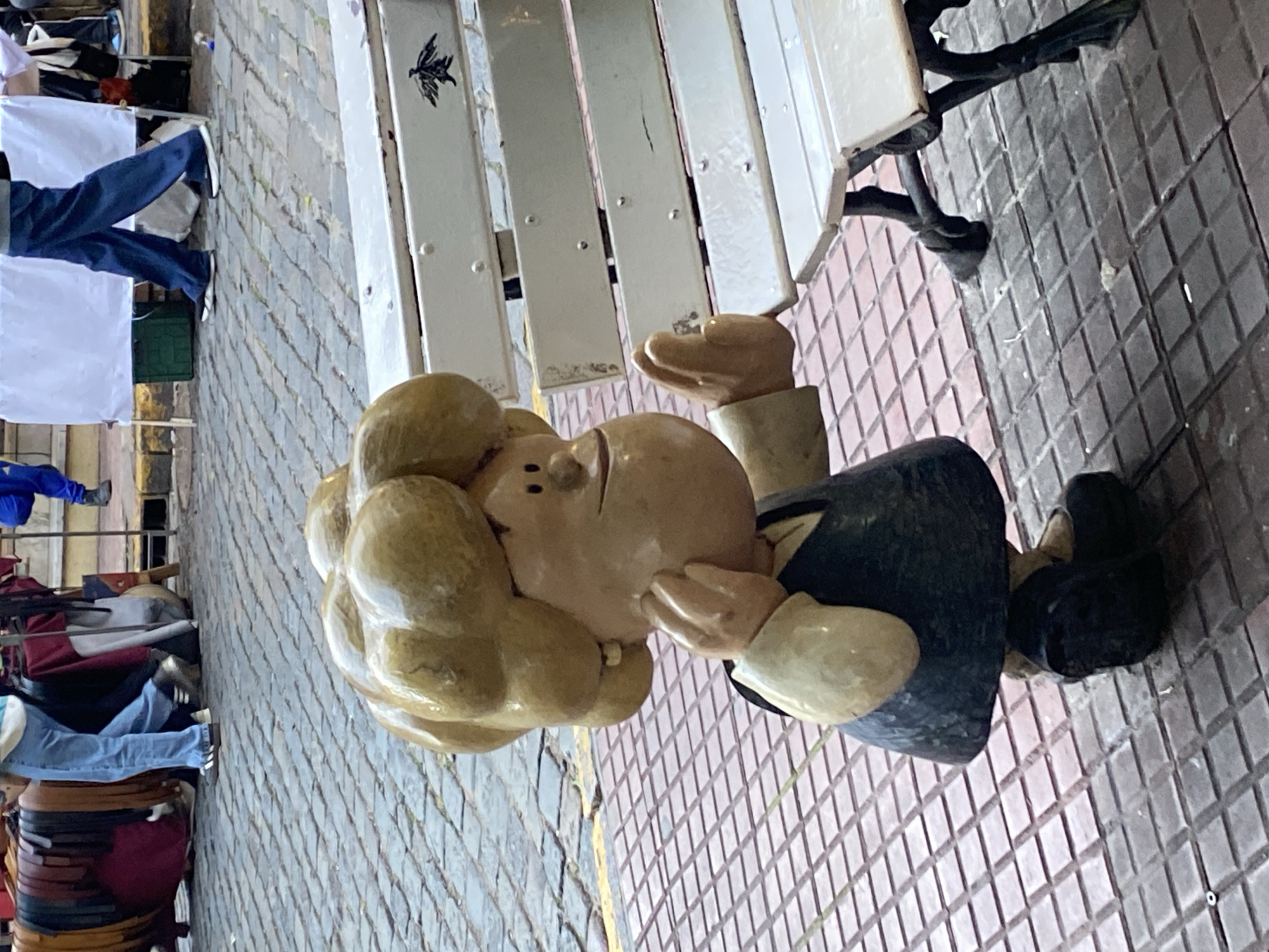 A sideways, medium shot depicts a wood statue of the comic strip character, Snoopy, seated on a white park bench in Buenos Aires, Argentina. Snoopy has blonde hair and is wearing a dark suit. The cobblestone street is visible to the left and right. People and market tables are also visible on the left side of the frame.