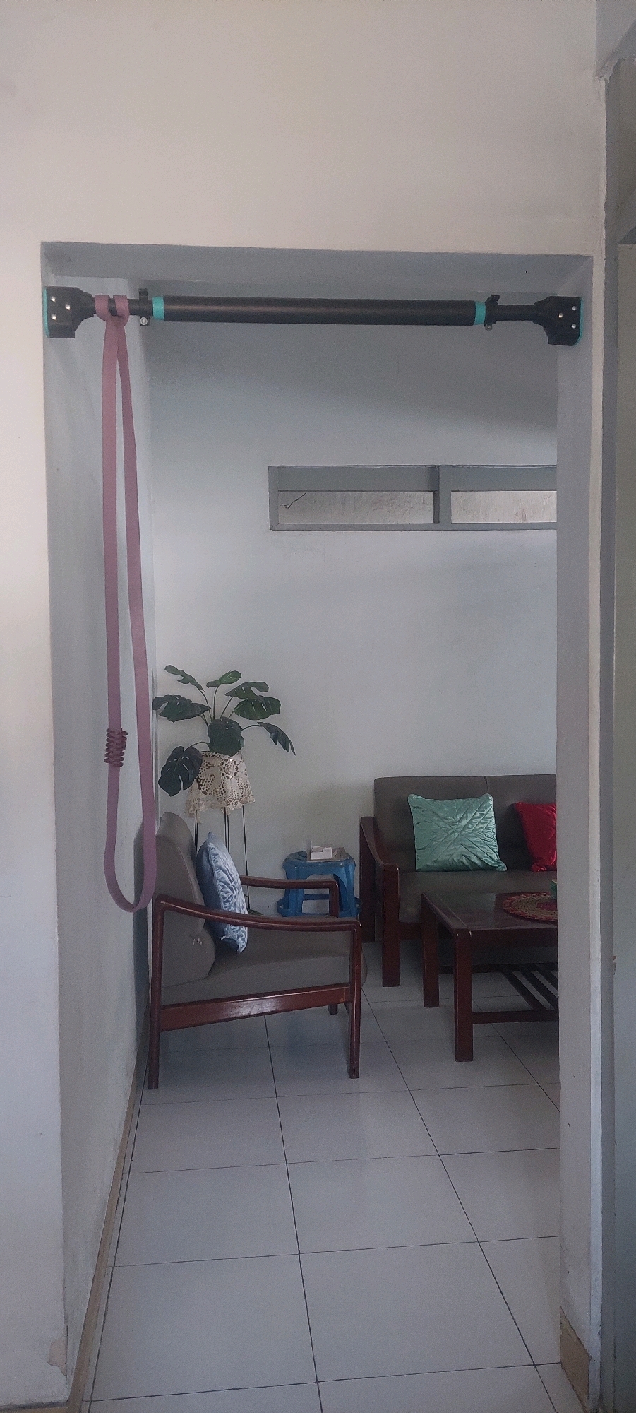 The image shows a doorway leading into a room with tiled floors and white walls. A dark-colored pull-up bar is mounted across the top of the doorway, with a long pink resistance band hanging from the left side. To the left of the doorway, a portion of a white wall is visible.
Inside the room, a single wooden chair with a blue pillow is positioned in the foreground on the left. Behind the chair, a potted plant with large green leaves sits on a stand covered with a doily. To the right of the plant, a small blue stool with items on top is visible. Further to the right, a wooden sofa with a light green and a red pillow is placed. In front of the sofa is a dark wooden coffee table. A rectangular window or vent is situated high on the back wall. The overall scene suggests a domestic indoor setting, likely a living area or hallway.