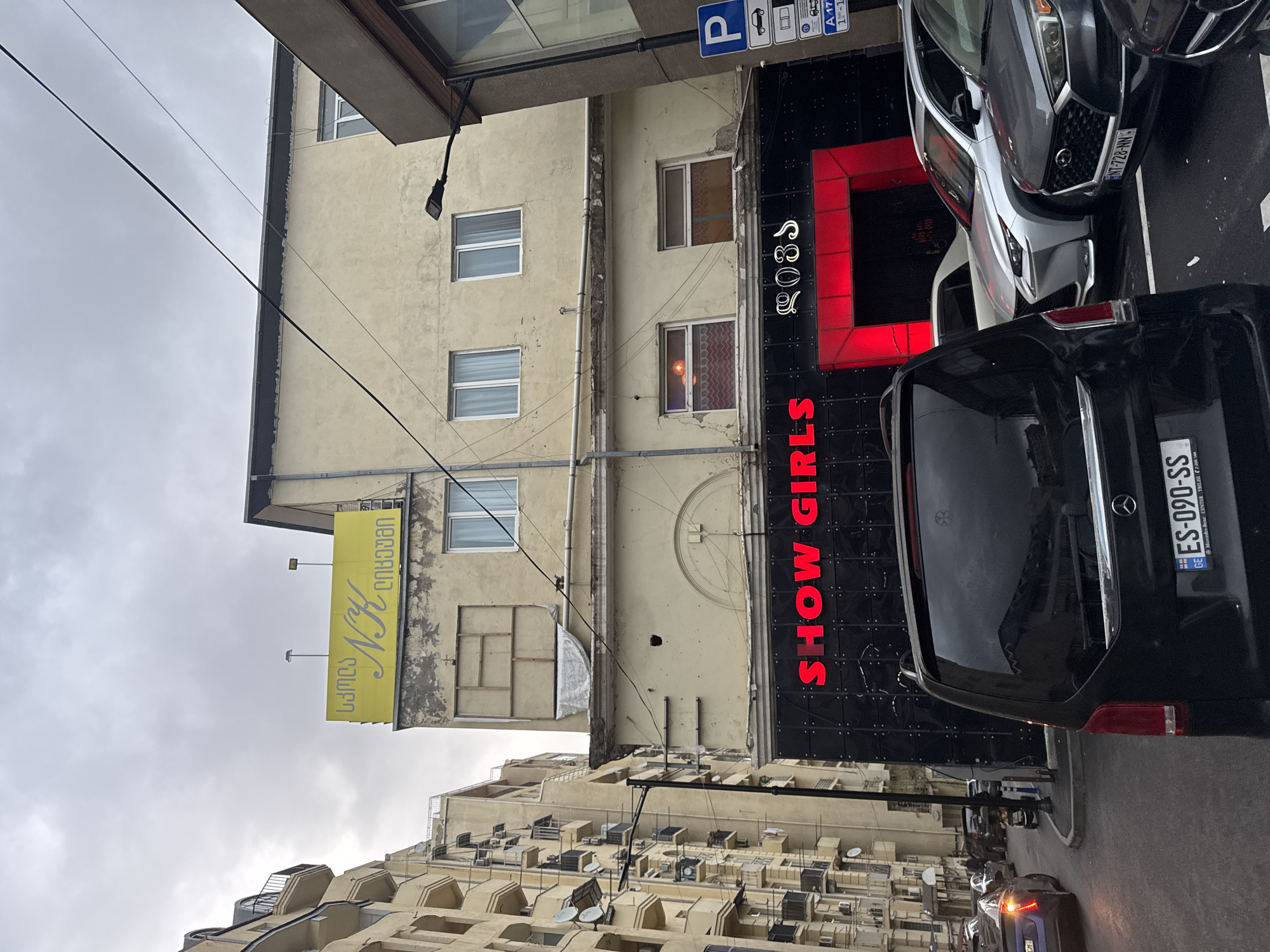 An urban street scene in Tbilisi, Georgia, features buildings, parked vehicles, and overhead utility lines under an overcast sky.
The prominent structure on the right side of the image is a commercial building with a dark facade displaying a vertical red neon sign that reads "SHOW GIRLS." Above this, another red neon sign spells out "RŌSS". This suggests a nightlife or entertainment establishment.
Adjacent to this, on the left, is a lighter-colored, somewhat weathered multi-story building with several windows. On its upper left side, a vertical yellow sign is affixed, featuring text in Georgian script. The visible text on this sign, as deciphered, is "სამღებრო NK დიზაინი" (Samghebro NK Dizaini), translating to "Painting NK Design."
Several vehicles are parked along the street. A black Mercedes-Benz van is prominently positioned in front of the "SHOW GIRLS" facade, displaying a Georgian license plate "GE ES-090-SS." Behind it, a silver or grey car with an Armenian license plate "AM-728-NN" is partially visible. Other cars can be seen further down the street.
Overhead, numerous electrical wires crisscross against the dull grey sky. A blue parking sign with a white "P" symbol and various vehicle pictograms (car, truck, bus) along with numbers like "A 17" and "1-1" is visible near the top center. The overall impression is that of a bustling city street during daytime, likely experiencing typical urban activity.