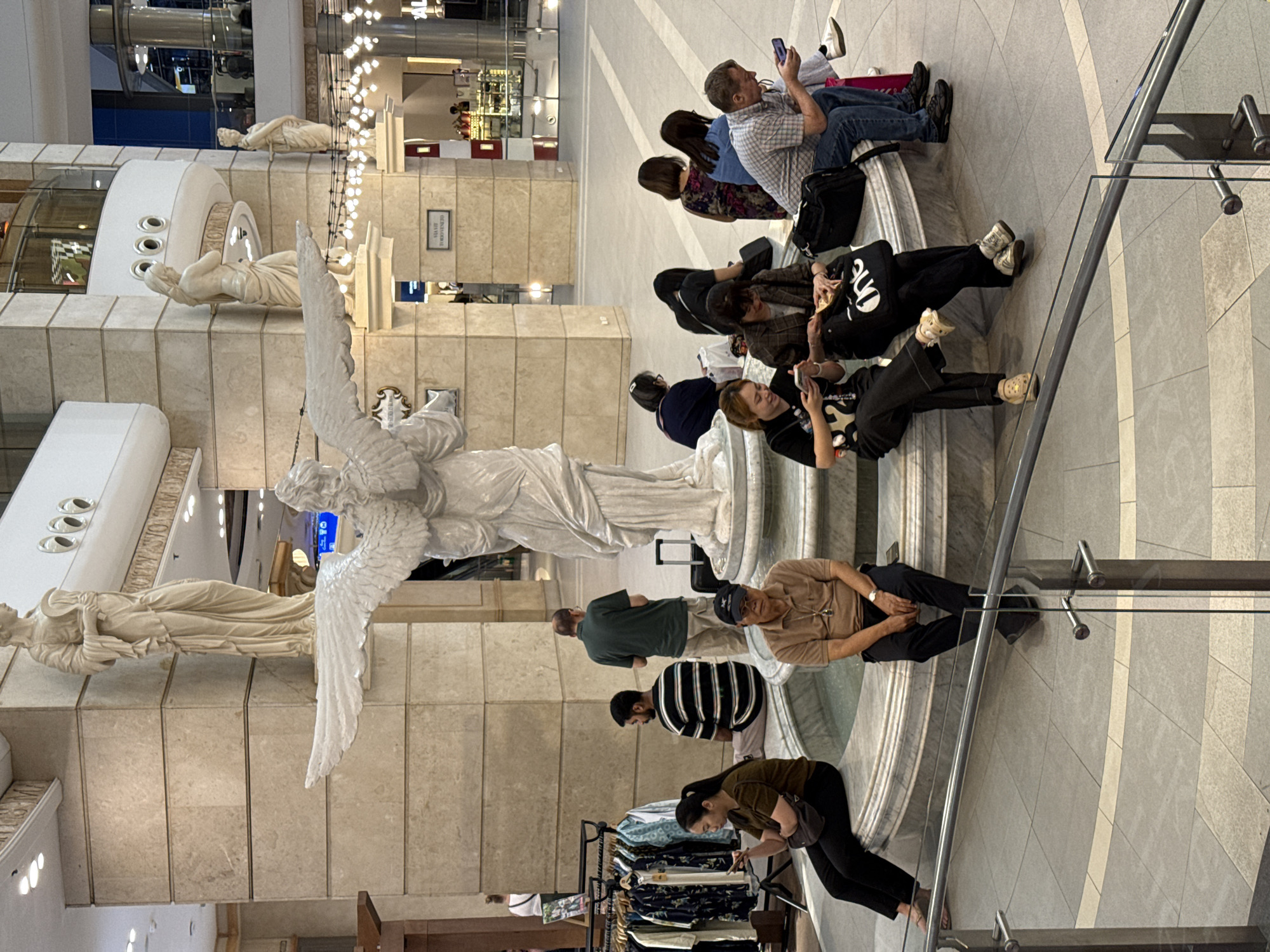 The image shows an interior scene in what appears to be a large, ornate shopping mall, likely located in Bangkok, Thailand, given the architectural style and general ambiance. A large marble fountain featuring a prominent sculpture of an angel with outstretched wings serves as a focal point. Several people are resting on the edge of the fountain, engaging in various activities such as using mobile phones, talking, or simply relaxing. A clothing rack with items for sale is visible near the fountain. The flooring is tiled, and the architecture features marble columns and decorative elements. Overhead, there are intricate lighting fixtures and balconies with additional statues. The scene depicts a blend of art, commerce, and relaxation within a public space.