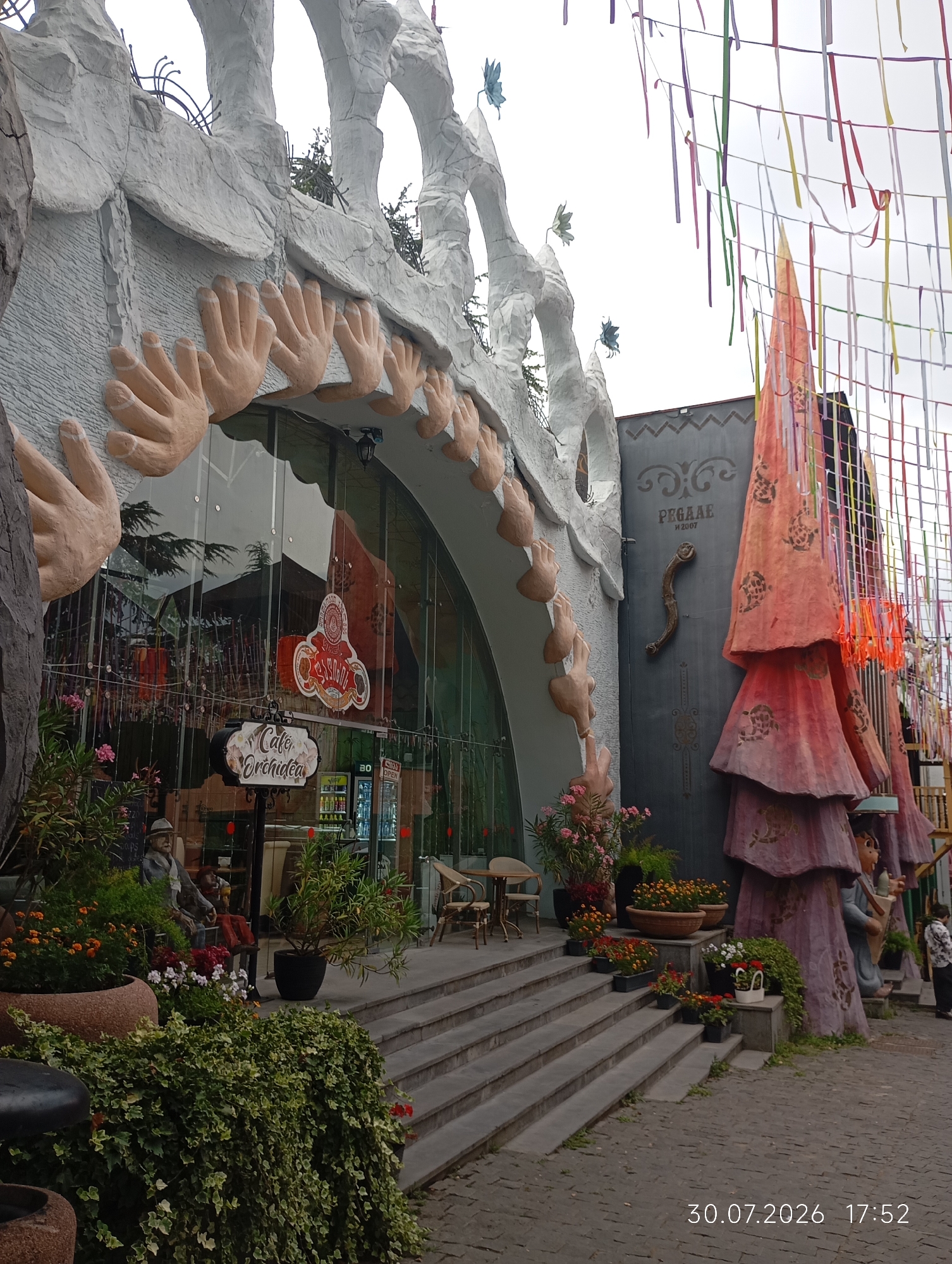 The image depicts the exterior of a uniquely designed building, likely a cafe or restaurant, in Tbilisi, Georgia. The architecture features large, stylized hands adorning the upper facade and an arched entrance with a glass-fronted interior. Colorful streamers hang overhead, creating a festive atmosphere. In front of the building, steps lead up to the entrance, flanked by potted plants and decorative figures. To the right, a tall, cone-shaped sculpture with a playful design stands prominently. A woman in a patterned shirt is visible in the lower right corner, walking away from the camera. The ground is paved with cobblestones. The timestamp "30.07.2026 17:52" is visible in the lower right corner, indicating the date and time the photograph was taken.