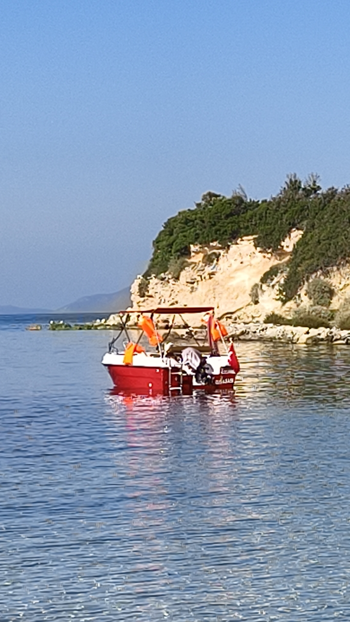 A small red boat with an orange canopy is floating on clear blue water. The boat has several orange life jackets secured to its exterior. A Turkish flag is visible at the stern of the boat. In the background, a rocky cliff covered in green foliage rises from the water's edge. The sky is a clear, bright blue. The scene appears to be a calm day in a coastal area, likely the Aegean Sea, near Kuşadası, Turkey, given the Turkish flag.