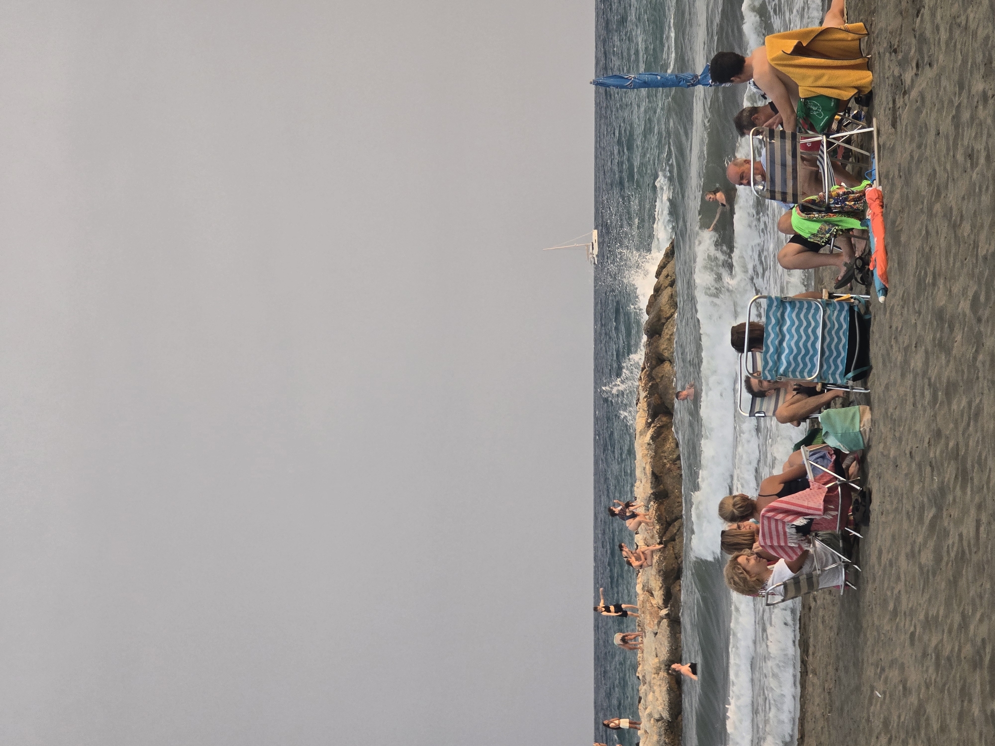 This image depicts a beach scene at sunset, with the city of Málaga, Spain, in the background. The foreground is a sandy beach with several groups of people relaxing in beach chairs. In the middle ground, waves crash against a rocky jetty where several individuals are standing and interacting. Further out in the sea, a sailboat is visible. The sky is overcast, contributing to a soft, diffused light.