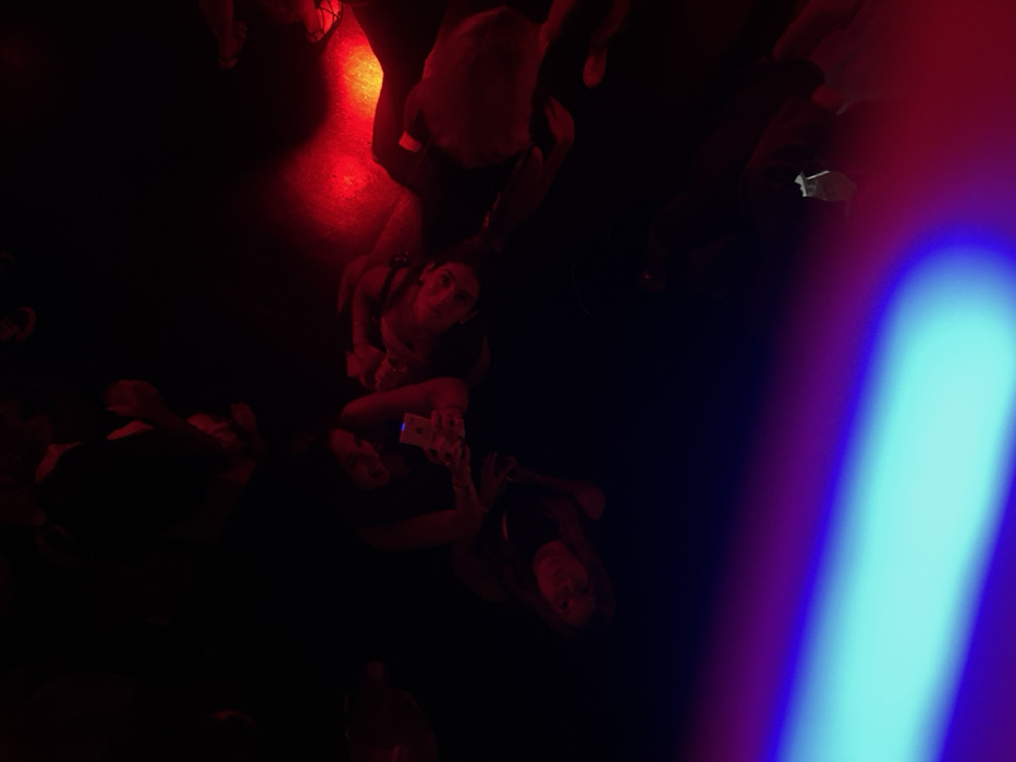 An overhead, low-light image depicts an interior nightlife venue in Paris, France. The environment is predominantly dark, illuminated by strong artificial lighting, including intense red light across the mid-right and upper sections, and a prominent, broad beam of blue-to-purple light extending diagonally from the lower-left corner.
Several individuals are present. In the mid-ground, three primary figures are discernible. One central person is seated or crouched, actively viewing a smartphone, the screen of which emits a distinct white-blue light onto their face and hands. To their right, a second individual is partially visible, appearing to be seated or reclining, with their face angled upwards, illuminated by the red light. A third person is partially visible to the left of the smartphone user. Additional indistinct, silhouetted figures are visible in the darker background and upper frame, indicating a crowd. No specific objects beyond the smartphone are clearly identifiable. The scene conveys a social gathering within a club or similar entertainment establishment.