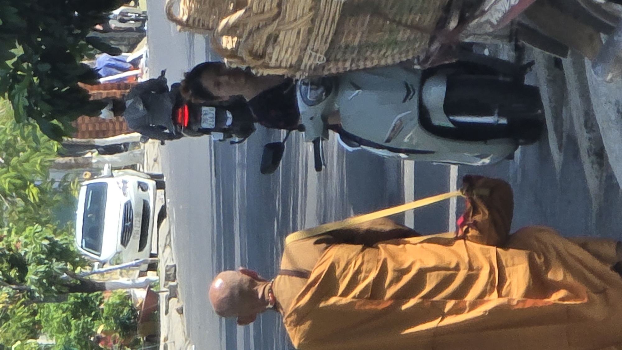 The image shows a street scene in Bắc Nha Trang, Vietnam. A Buddhist monk with a shaved head, wearing a saffron robe, is walking in the foreground. A woman is riding a light green scooter with a basket on the front. A white vehicle is parked on the left side of the road. The road is paved with white lines marking lanes. Trees and greenery are visible on the left side of the image.