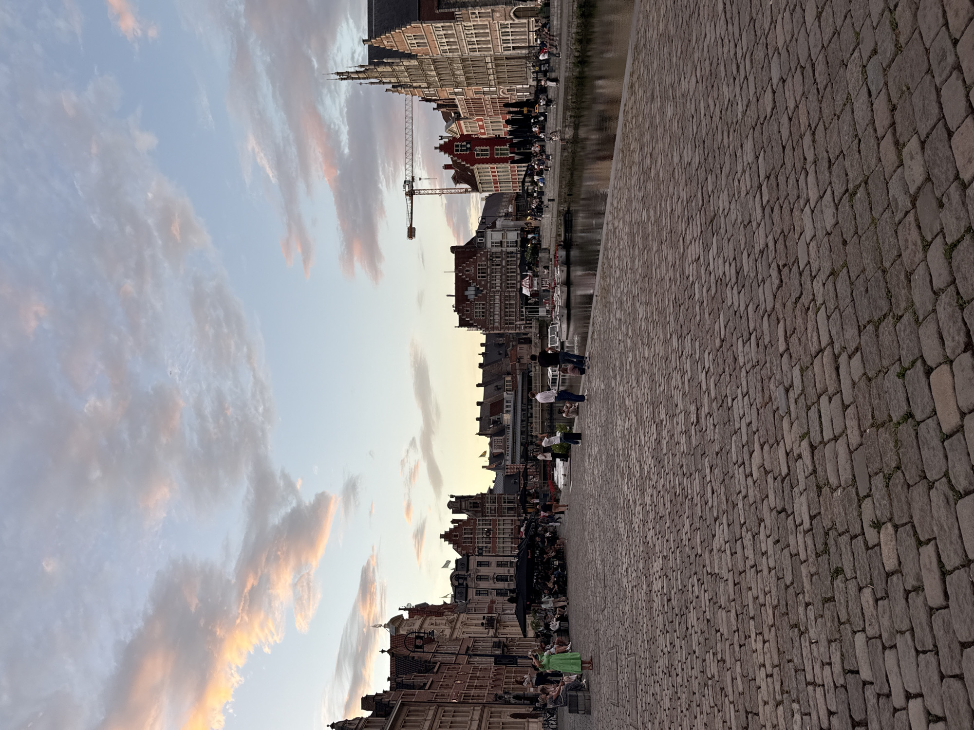 The image depicts a twilight scene along a canal in Ghent, Belgium, likely the Graslei or Korenlei. The foreground is dominated by a wide, uneven cobblestone pavement leading towards the water. On the far side of the canal, a row of historic, red-brick buildings with steeply pitched roofs and ornate gables lines the waterfront. Several people are visible on the pavement and along the canal edge. On the left, a woman in a green dress stands out amongst a group of seated individuals. Further along the canal, more people are seated, some at tables under black umbrellas, suggesting outdoor dining. Boats are moored along the quay. In the background, a construction crane is prominent, contrasting with the historic architecture. The sky above is a soft blend of pale blue and pink hues, with scattered clouds tinged by the setting sun. The overall atmosphere is calm and pleasant, with people enjoying the evening by the water.