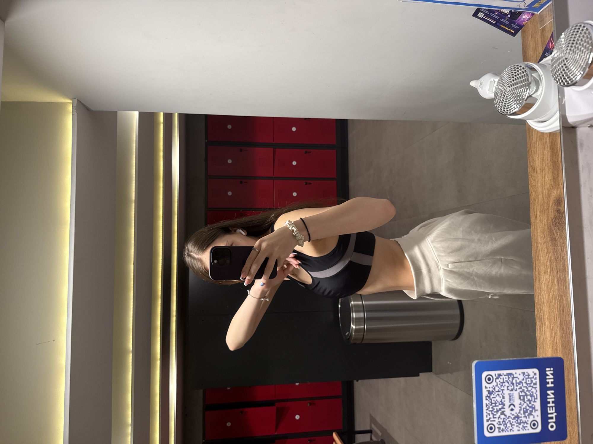 A young female, likely in her late teens or early twenties, is captured in a mirror selfie within a changing room or locker room in Sofia, Bulgaria. She has long, straight dark brown hair and is wearing a black sports bra with gray and white panels and light-colored sweatpants. On her left wrist, she wears a white scrunchie and a dark beaded bracelet, while her right wrist displays a silver-colored bracelet with a small charm and a ring on her ring finger. White wireless earbuds are visible in both ears, and her fingernails are painted white. She holds a dark-colored smartphone, likely an iPhone Pro Max, obscuring her face, as she photographs her reflection.
The background shows rows of bright red lockers with silver handles, consistent with a gym or fitness facility. A silver cylindrical trash can stands on the left. The ceiling features multiple parallel recessed linear light fixtures emitting a warm glow. In the foreground, a wooden counter is visible, on which a blue square sign with a white border sits. The sign displays a QR code, the text "next level," and "ОЦЕНИ НИ!" (Bulgarian for "RATE US!"), indicating the location is in Bulgaria. To the right of the sign, two white devices with chrome mesh tops, resembling hairdryers or personal grooming appliances, are positioned on the counter. The image is a reflection, captured in a mirror with a gray tiled floor visible below.