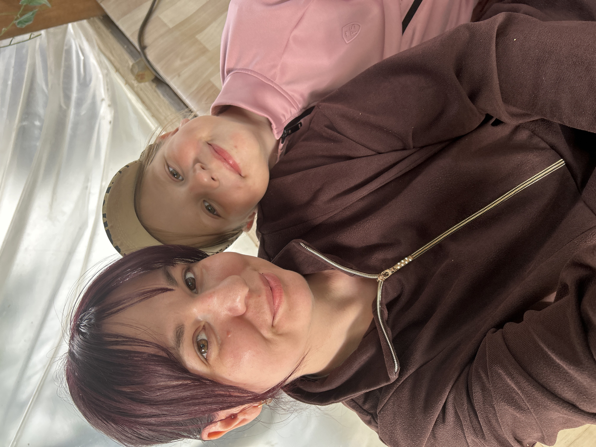 A close-up, upward-angle, self-portrait photograph taken indoors in the city of Фоки, Russia, features an adult female and a young child. The adult female is positioned in the foreground, left of center, with dark reddish-brown hair styled with bangs and brown eyes. She has a light complexion with visible redness on her cheeks and a small gold earring with a dangling element in her left ear. She is wearing a dark brown, textured zippered top with a gold-colored zipper featuring a pull tab adorned with small, pearl-like beads. To her right and slightly behind her, a young child, possibly female, rests their head on the adult's left shoulder. The child has light-colored hair partially obscured by a light-colored, possibly beige or camouflage-patterned, baseball cap, and light-colored eyes with rosy cheeks. The child is wearing a pink athletic-style zippered top with a black zipper and a small embroidered crest on the left chest. Both individuals are looking directly at the camera with faint smiles. The background consists primarily of draped, translucent white plastic sheeting or tarpaulin in the upper section, creating a corrugated texture, suggesting a temporary or greenhouse-like structure. A section of light-colored flooring or a wall panel is visible in the lower right. In the upper right corner, a wooden beam and a small green leafy plant are visible.