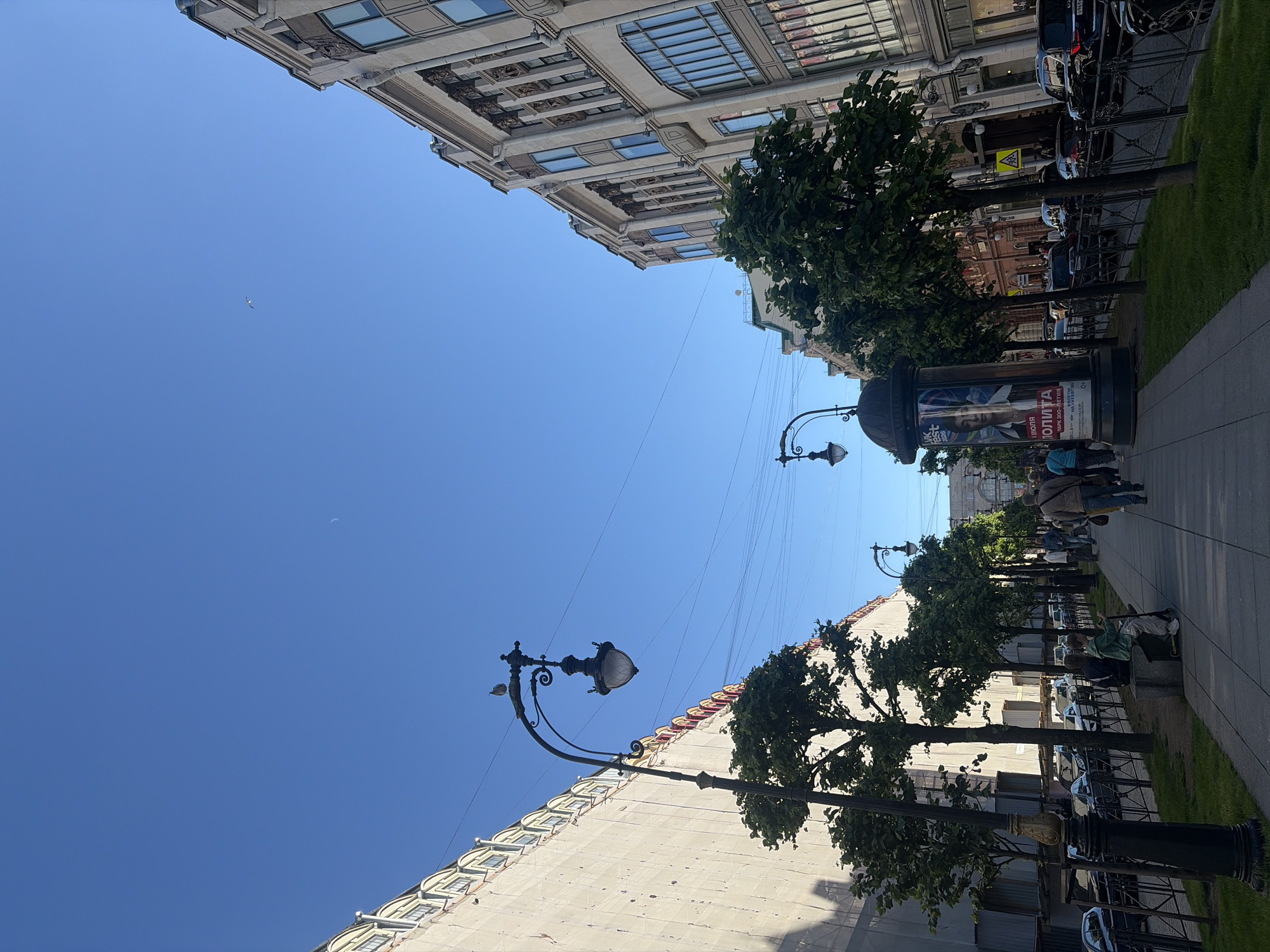 This is a street scene in Saint Petersburg, Russia, on a clear day with a blue sky. The perspective is looking down a tree-lined sidewalk. On the left, a large building with scaffolding is visible, with ornate yellow and red trim. On the right, a more detailed building with classical architecture and large windows stands.
Several people are walking on the sidewalk. Some are in the foreground, while others are further down the street. A prominent lamppost with a bird perched on it stands in the middle ground on the left. A large poster on a cylindrical display is also visible on the left, featuring a person's face and Russian text. Cars are parked along the street on the left.
The sidewalk is paved and bordered by a black metal fence. Trees with green foliage line the sidewalk, casting shadows on the ground. The overall atmosphere appears to be one of a typical urban street in a historic European city.