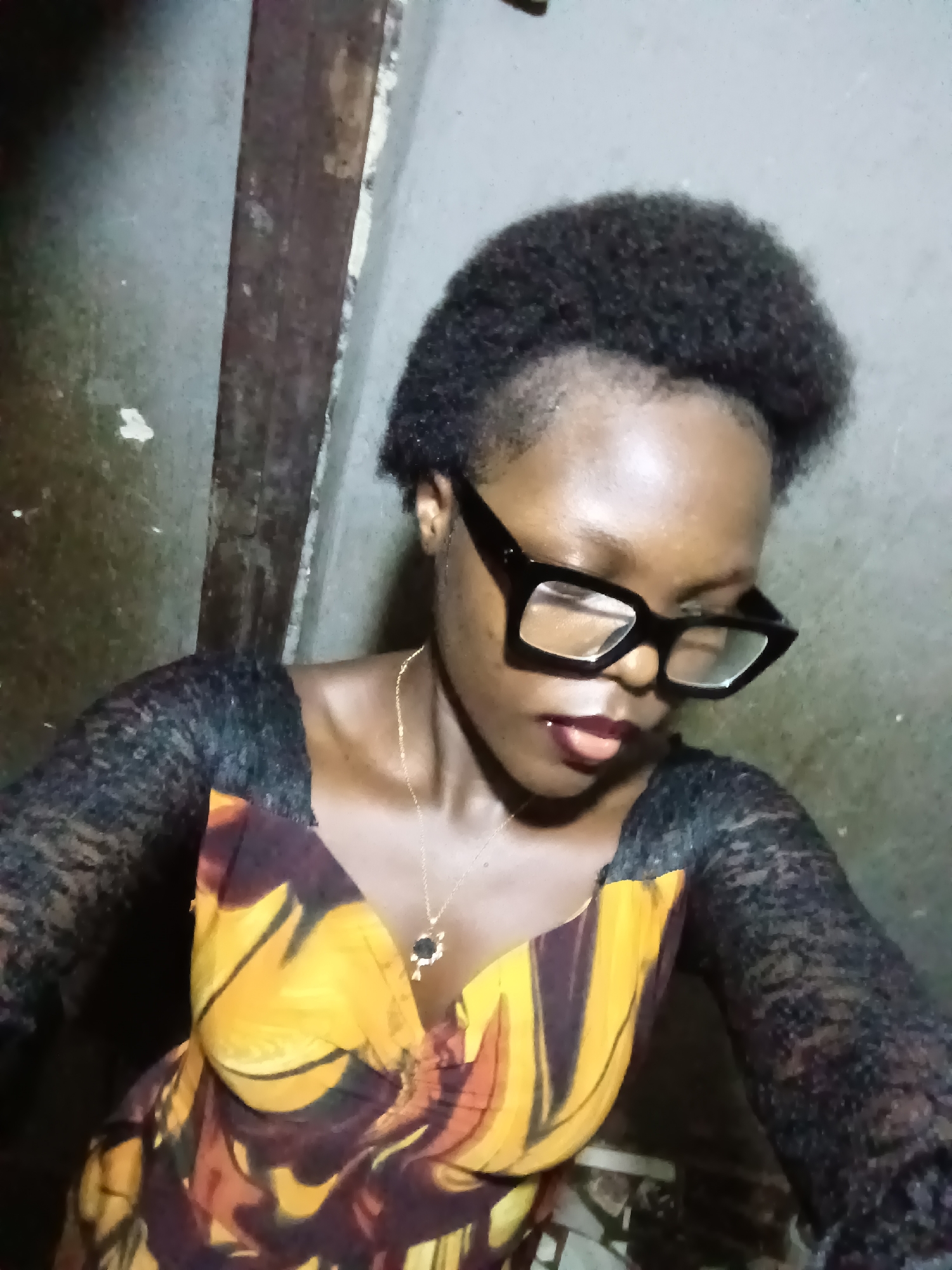 A Black woman with short, natural hair and large, black-rimmed glasses is shown from the shoulders up, looking down and to her right. She is wearing a yellow and brown patterned dress with sheer black sleeves. A thin gold chain with a dark pendant is visible around her neck. The background is a textured, grey wall with a vertical wooden beam. The lighting is dim, suggesting an indoor setting, possibly a home in Onitsha, Nigeria.