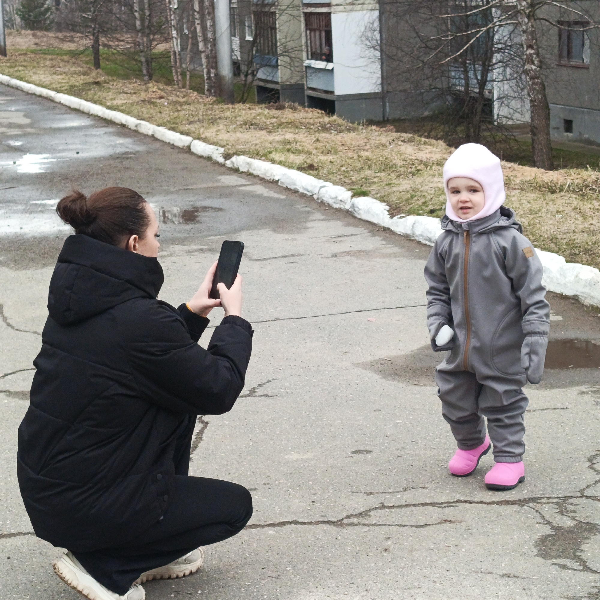 A woman is kneeling and taking a photograph with a black smartphone of a young child who is standing on a cracked asphalt road. The child is wearing a gray jumpsuit with a pink balaclava-style hat and pink rubber boots. The woman is wearing a black puffy jacket, black pants, and off-white sneakers. The backdrop includes a road bordered with a white curb and nondescript buildings with bare trees and patches of dormant grass. The location is Марковский, Russia.