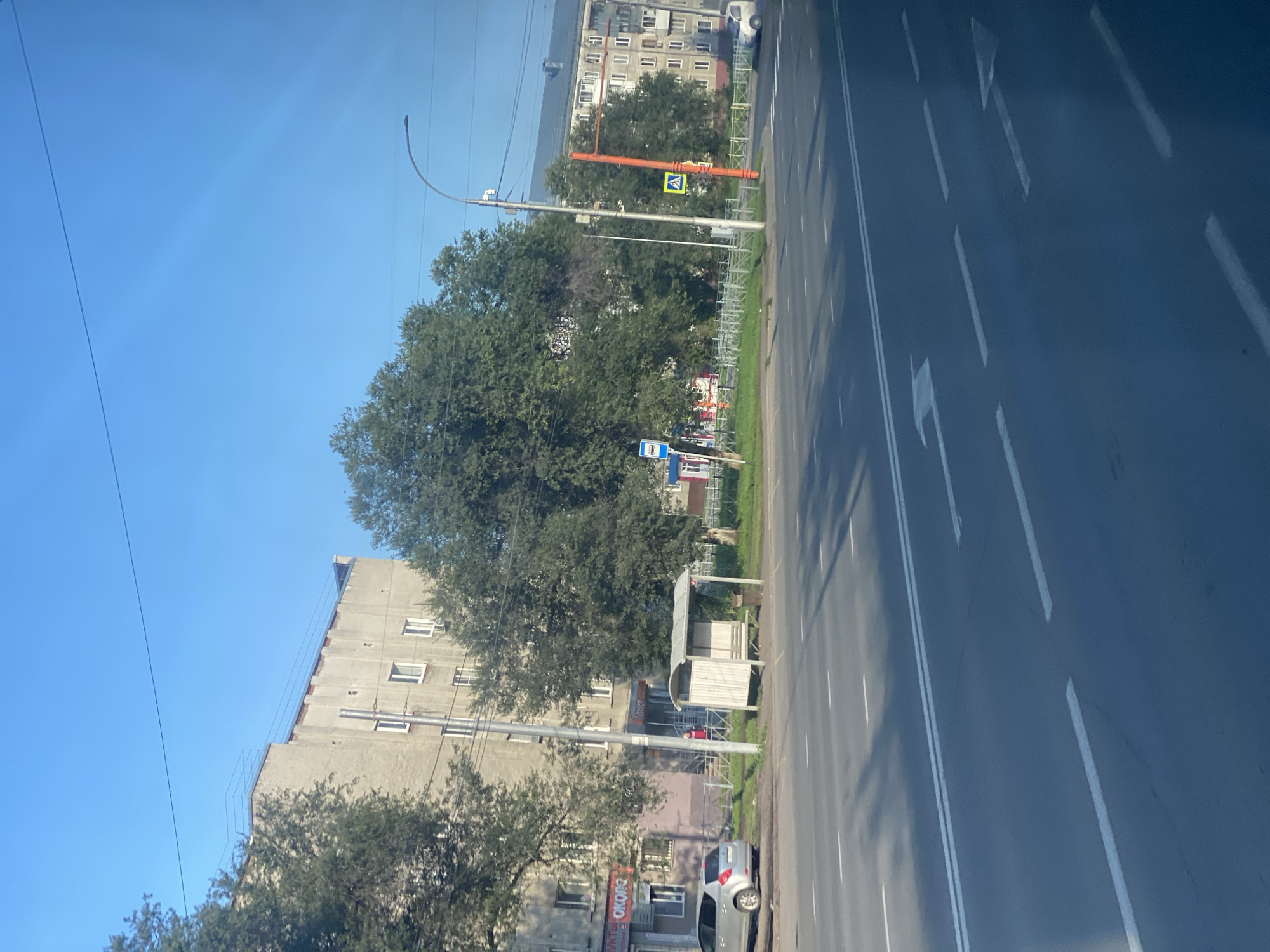 A street scene in Kemerovo, Russia, is depicted under a clear blue sky. The image captures a multi-lane asphalt road with white lane markings and directional arrows. On the left side of the road, a silver car is parked on the shoulder, adjacent to a building with shops at ground level. A bus stop shelter with a bench is also visible near the building. Tall trees line the sidewalk in front of the building. Further down the street, a modern street lamp with multiple fixtures stands prominently. The background features apartment buildings, with balconies and windows visible. The overall impression is of a typical urban street during daylight hours.