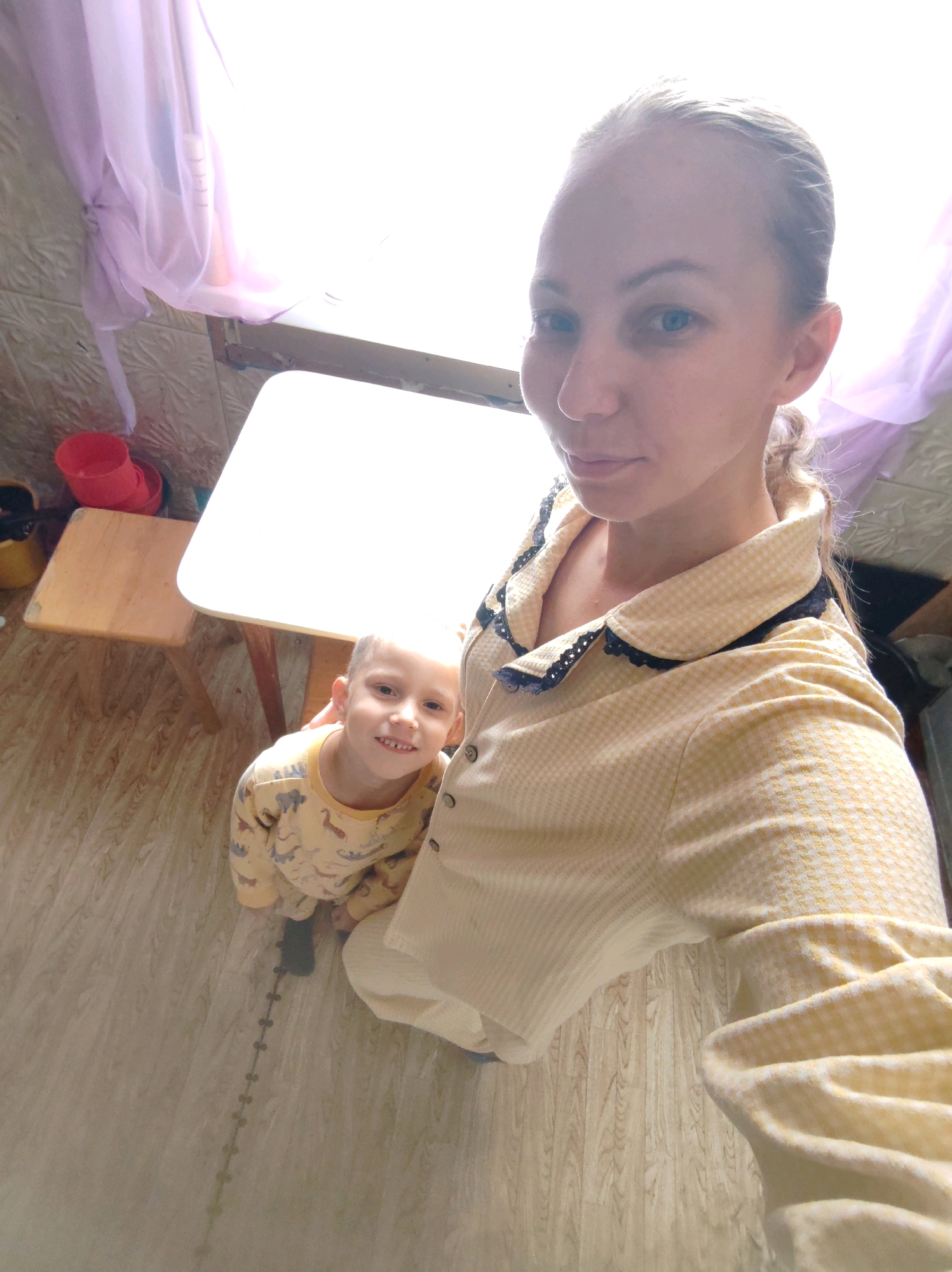 In the city of Chukhlem, Russia, a selfie is taken by a woman with blonde hair pulled back, wearing a yellow checkered pajama top with black trim. She is positioned in the foreground, looking at the camera with a slight smile. To her right and slightly in front is a young boy, also in yellow pajamas with a dinosaur print. He is smiling broadly at the camera. They are indoors, with a white table in front of them. To the left of the table, a small wooden stool is visible, with two red plastic buckets on top of it. The floor is a light wood laminate. A sheer purple curtain is draped over a window in the background, creating a bright, washed-out effect. The overall setting appears to be a domestic interior.