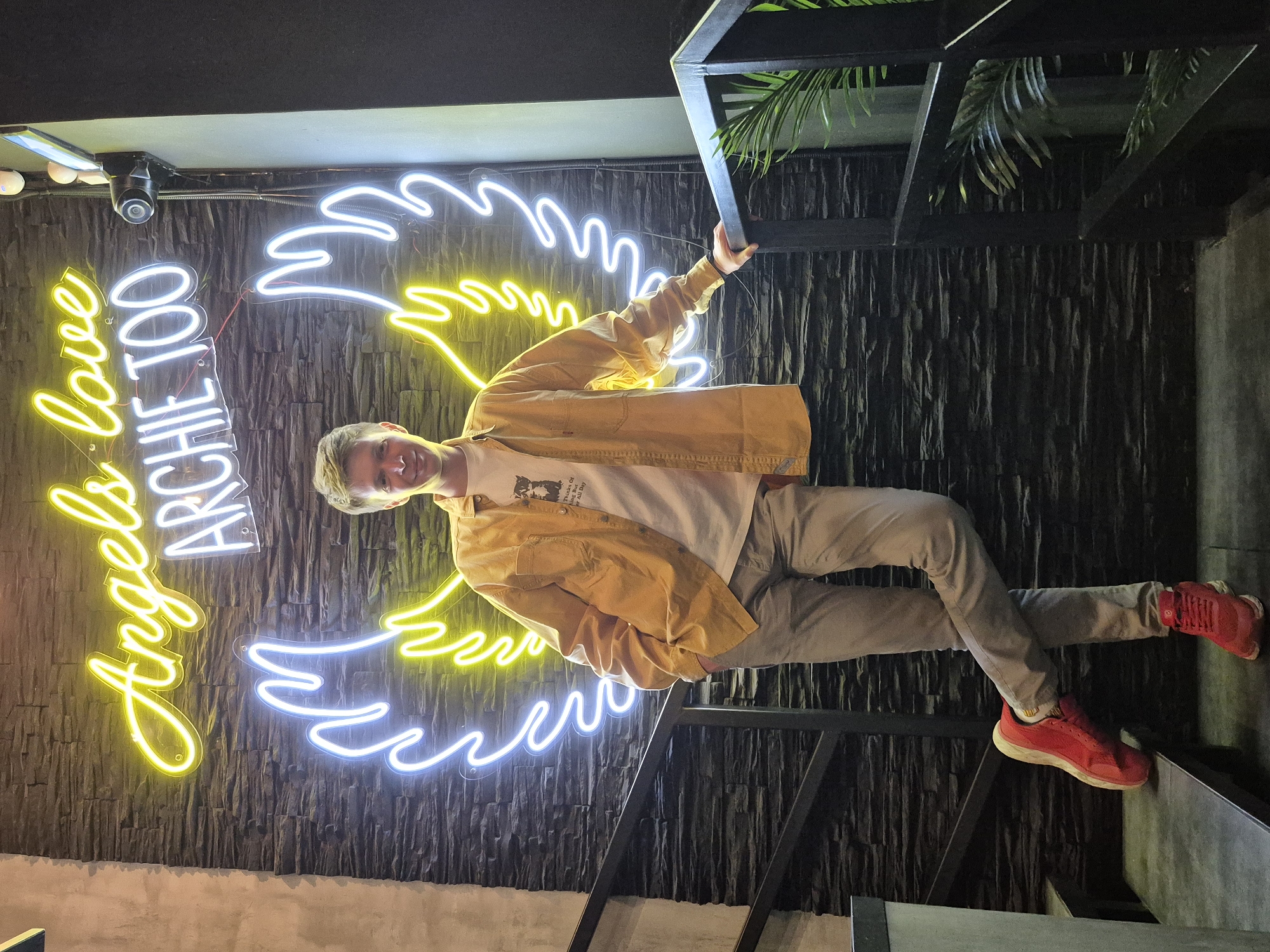A young Caucasian male with light brown hair and a clean-shaven face stands in front of a dark stone wall with a neon sign that reads "Angels love Archie too" in yellow and white, and a pair of angel wings outlined in blue and yellow. He is wearing a mustard yellow long-sleeved shirt over a white t-shirt with a graphic print, light grey trousers, and red sneakers. The male has his left leg crossed over his right, and his left hand is in his pocket. He is positioned on a dark grey staircase with a black metal railing. To the right of the male, a tropical plant is visible behind the railing. The overall lighting suggests an indoor setting with artificial lighting. The image was taken in Izhevsk, Russia.