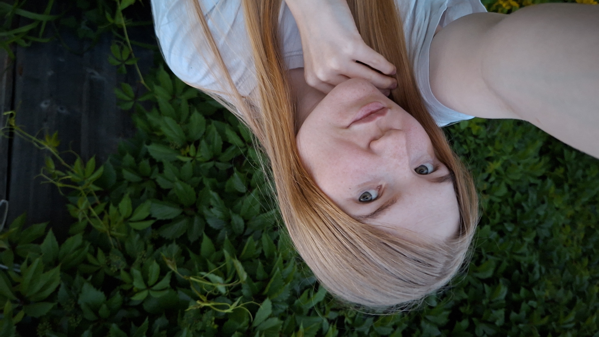 A young woman with long, blonde-red hair is taking a selfie outdoors. She is looking towards the camera with a slight smile and her left hand is resting on her chin. She is wearing a white t-shirt. Behind her is a dense wall of green leaves, likely part of a garden or natural foliage, and to the right, a dark wooden fence is visible. The lighting suggests it is late afternoon or early evening. The setting appears to be a residential area, possibly a backyard or garden in the village of Bolshoy Bukor, Russia.
