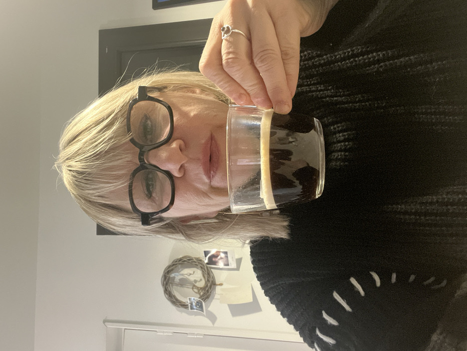 A woman with blonde hair and black glasses holds a clear glass filled with a dark beverage. She wears a black sweater with white stitch detailing. A silver ring is visible on her finger. The background shows a white wall adorned with a decorative wreath and a few small photos attached with tape. The image is turned sideways.