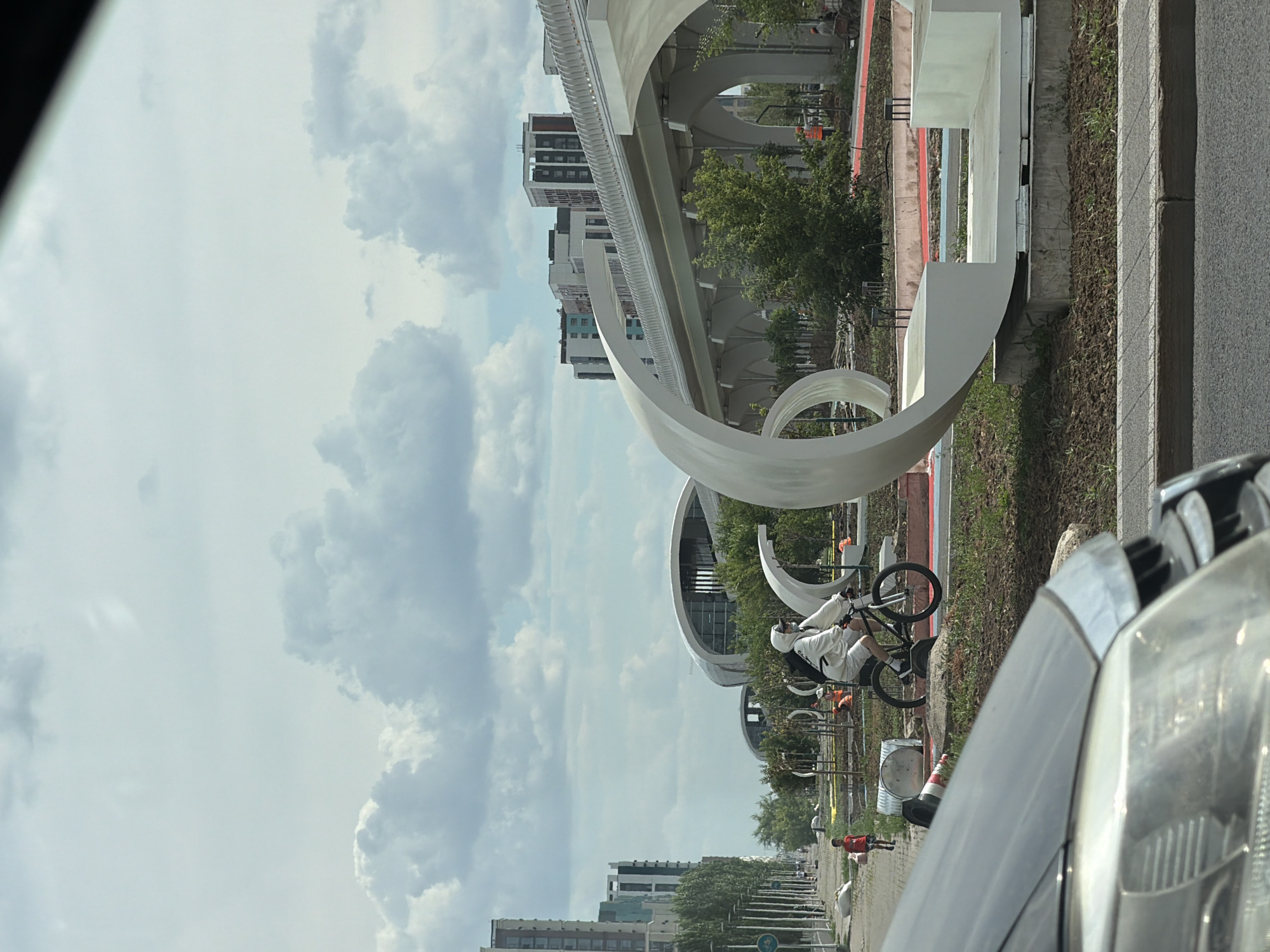The image depicts an outdoor scene with a modern architectural design. A person is riding a bicycle in the foreground, wearing a white outfit and a helmet. The bicycle is black and appears to be a BMX bike. Behind the cyclist, there's a large, abstract white sculpture with curved elements. The background features several modern buildings with multiple stories and windows, interspersed with green trees. There's also a pedestrian walkway and what appears to be a paved road or path. The sky is filled with clouds, suggesting it could be daytime with overcast weather. The image appears to have been taken from a vehicle, as the hood of a car is visible in the bottom right corner. The location is identified as Astana, Kazakhstan. The overall impression is one of contemporary urban development and recreation. There is no visible text in the image.