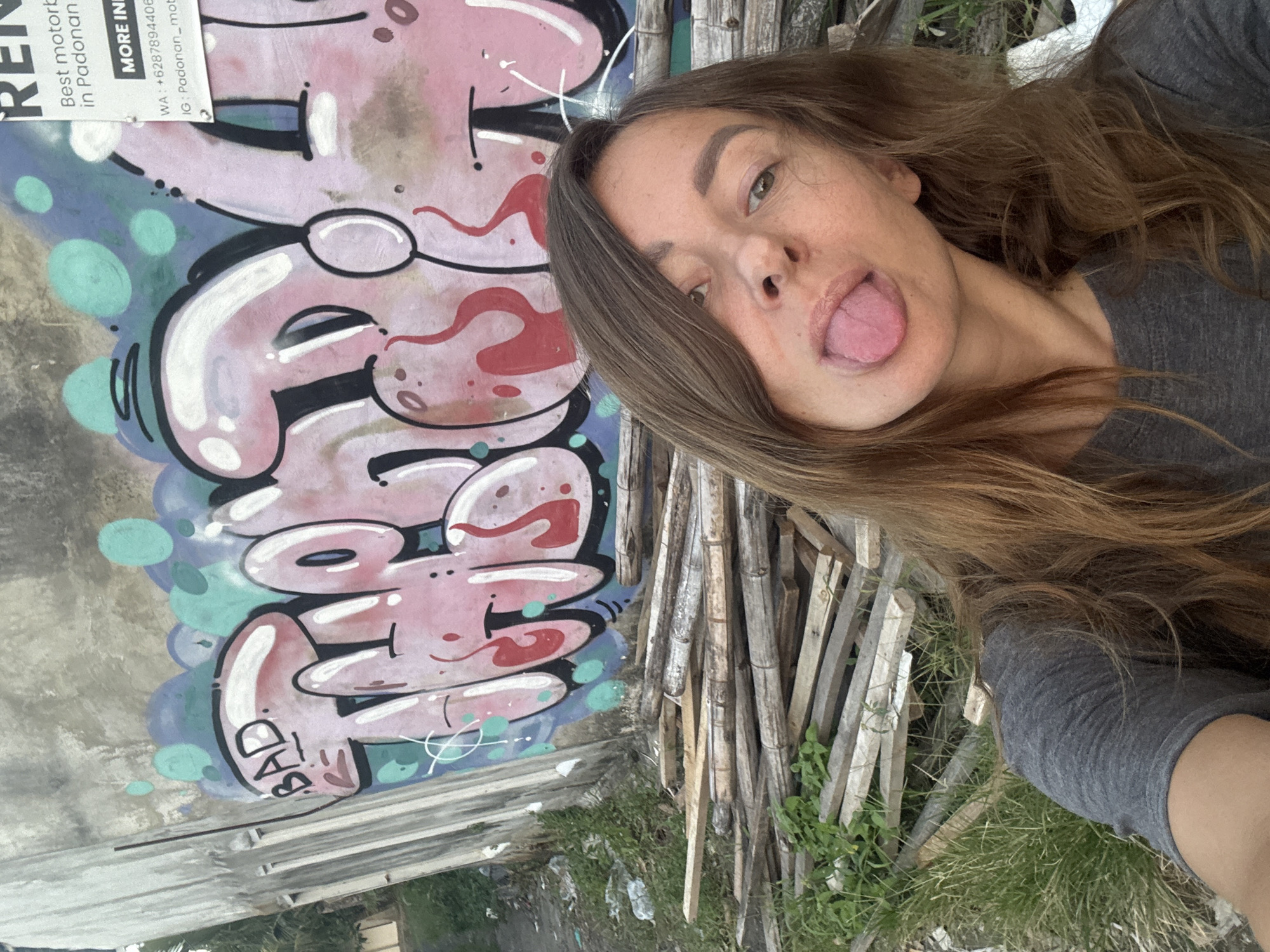 The image shows a woman sticking her tongue out in front of a wall with graffiti. The word "BAD" is visible on the wall. A sign promotes motorcycle rentals in Padonan, North Kuta, Indonesia, with contact information. Adjacent to the wall is a pile of weathered wooden stakes and grassy vegetation.