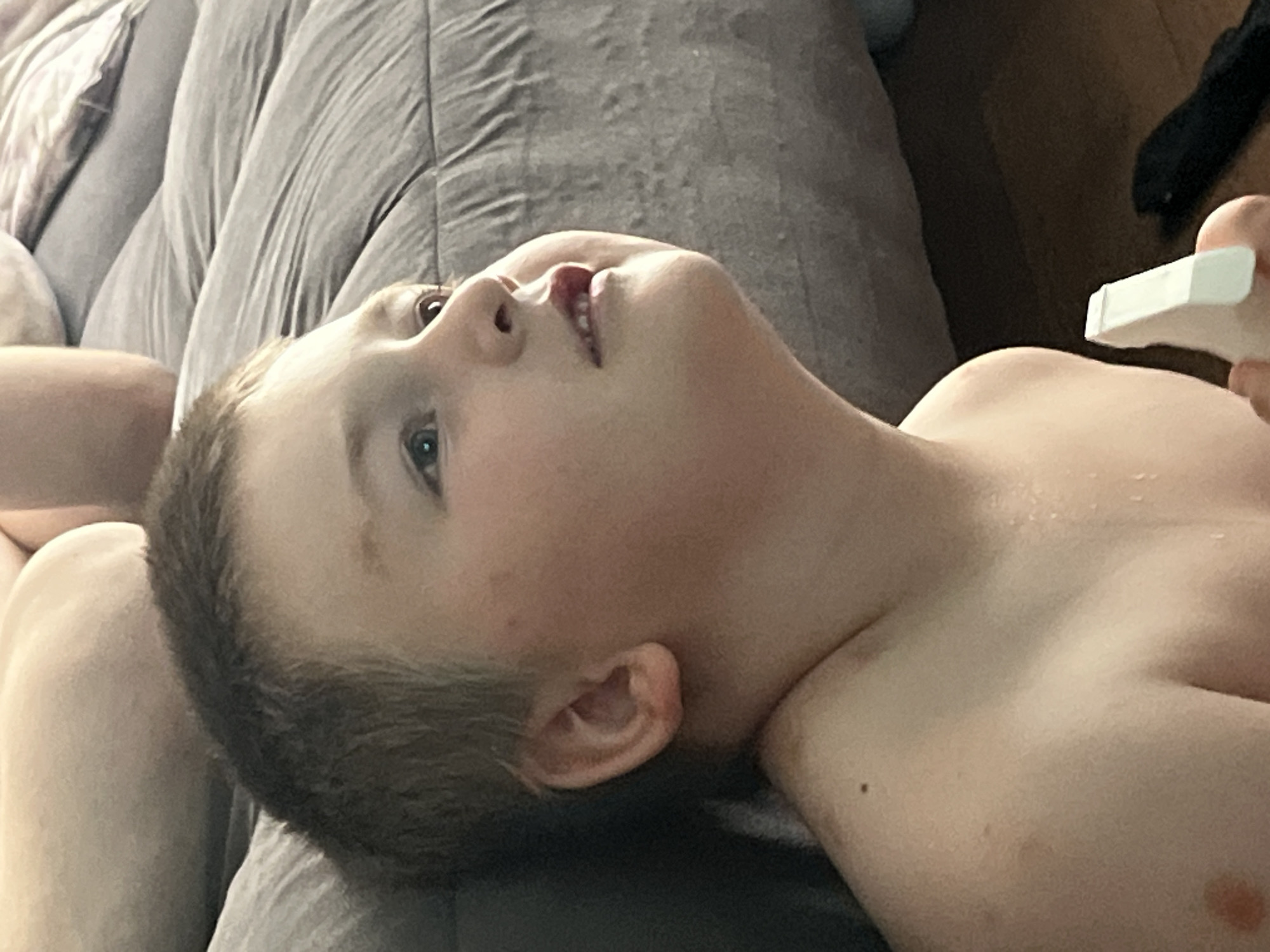 This media file shows a close-up, low-angle shot of a young boy lying on his back. He has fair skin, light brown hair, and blue eyes. His head is tilted back, and he is looking upwards. His mouth is slightly open, revealing his teeth. His right ear is visible. He is wearing a grey t-shirt, and appears to be on a couch or bed with a grey textured blanket. A person's hand is visible in the upper right corner, holding a white spray bottle. The lighting suggests it is daytime. The scene appears to be indoors. The location context is Chelyabinsk, Russia.