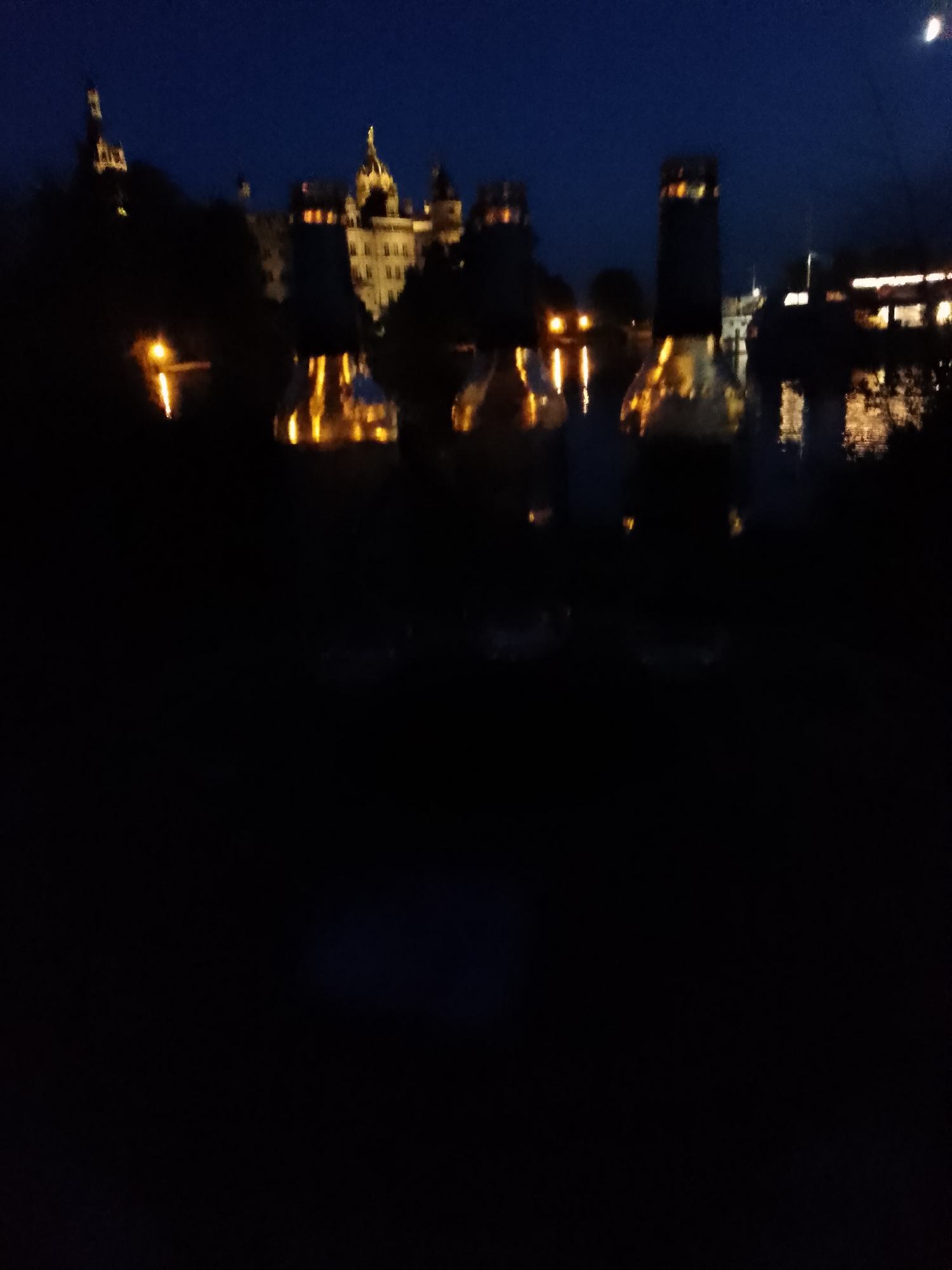 The image shows a blurry nighttime view of the Schwerin Castle in Schwerin, Germany, reflected in the water. The castle appears to be lit up, with lights visible in the windows and on the towers. The sky is dark blue, with a hint of a crescent moon in the upper right corner. The foreground is dark and out of focus, suggesting the photo was taken from a distance or in low light conditions. The overall impression is one of a serene and picturesque evening scene.