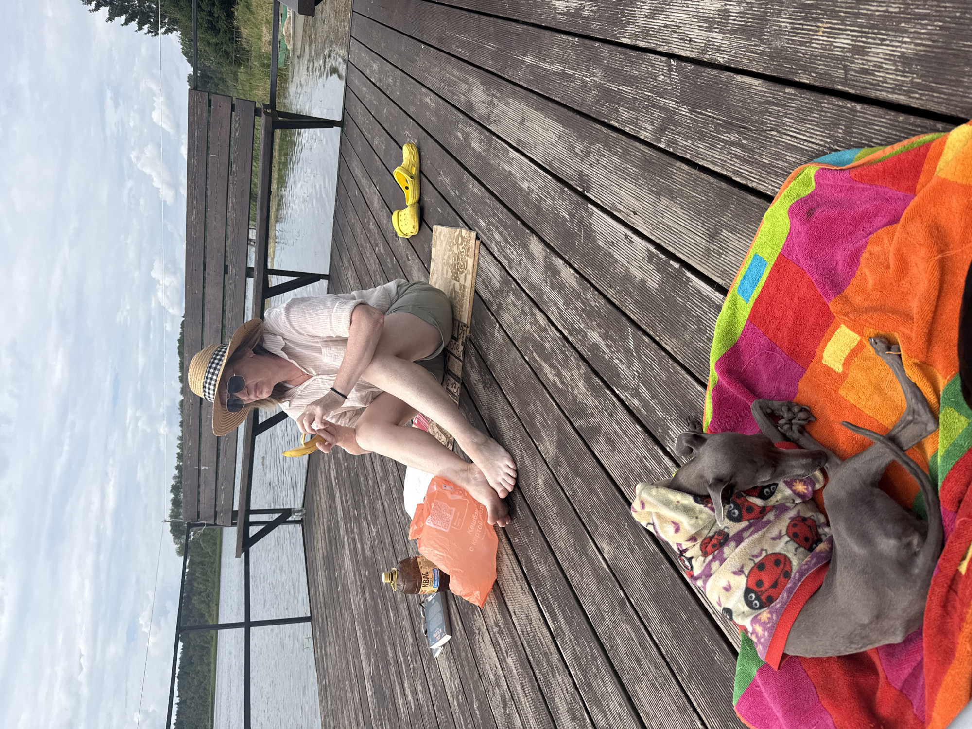 A woman wearing a straw hat, sunglasses, and a light-colored shirt is sitting on a wooden dock with a banana in her hands, appearing to be peeling it. Beside her are a bottle of Kvass, an orange plastic bag, and a book. A pair of yellow Crocs are a few feet away. In the foreground, a small grey dog wearing a ladybug-patterned sweater is asleep on a colorful blanket. The scene is set by a body of water with a wooden bench and trees in the background, under a cloudy sky. The location is the city of Chikino, Russia.