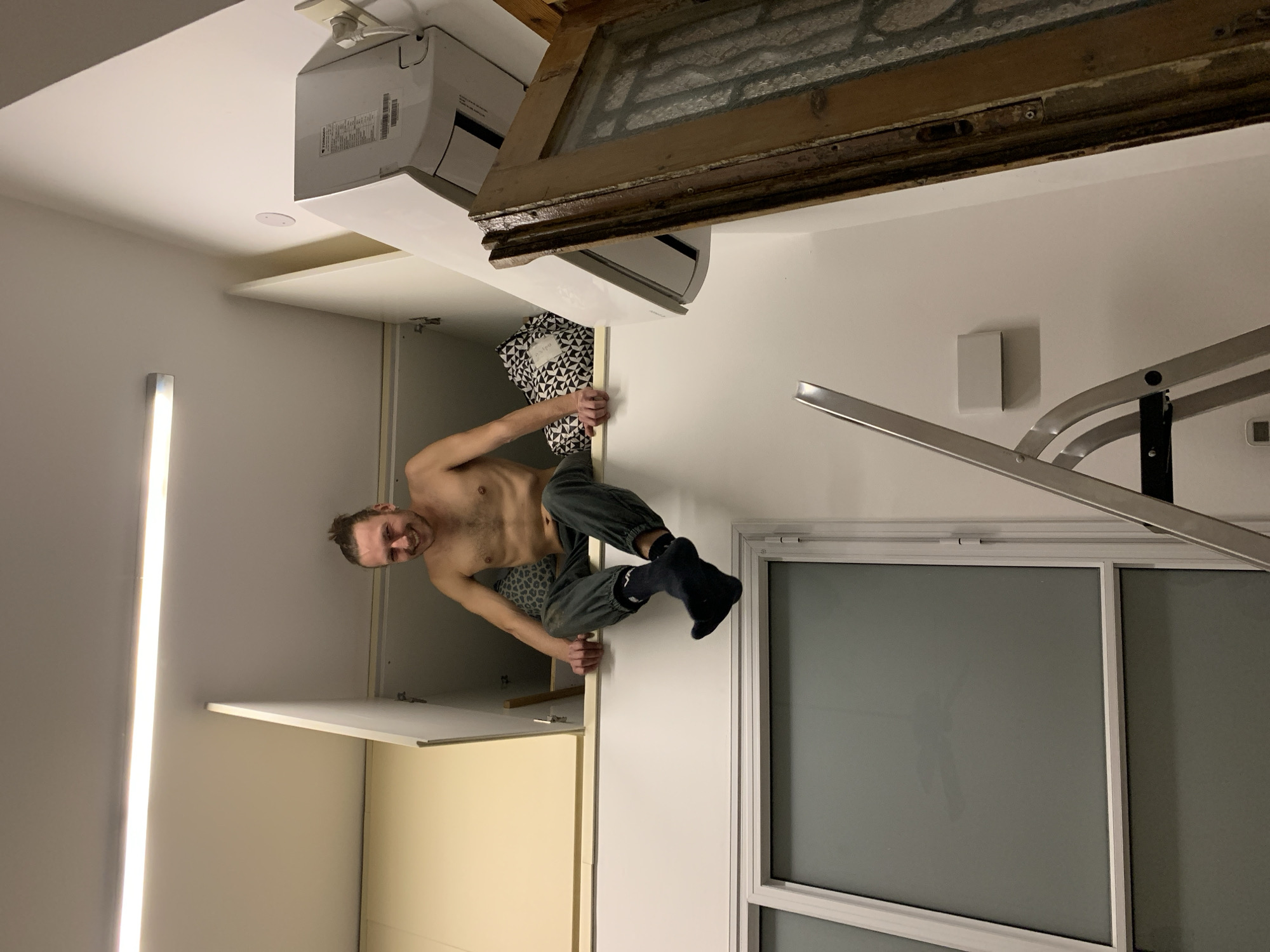 A man with a beard and mustache is shown in an indoor setting. He is climbing on top of a white cabinet and appears to be looking at the camera. The man is shirtless and wearing grey pants and dark socks. The room contains a window with a grey frosted glass pane and a drying rack. A light fixture is visible on the wall. Above the cabinet is an air conditioning unit and an old wooden frame, possibly a door or window frame. The location is unknown and there is no reference to Tel-Aviv or Israel.