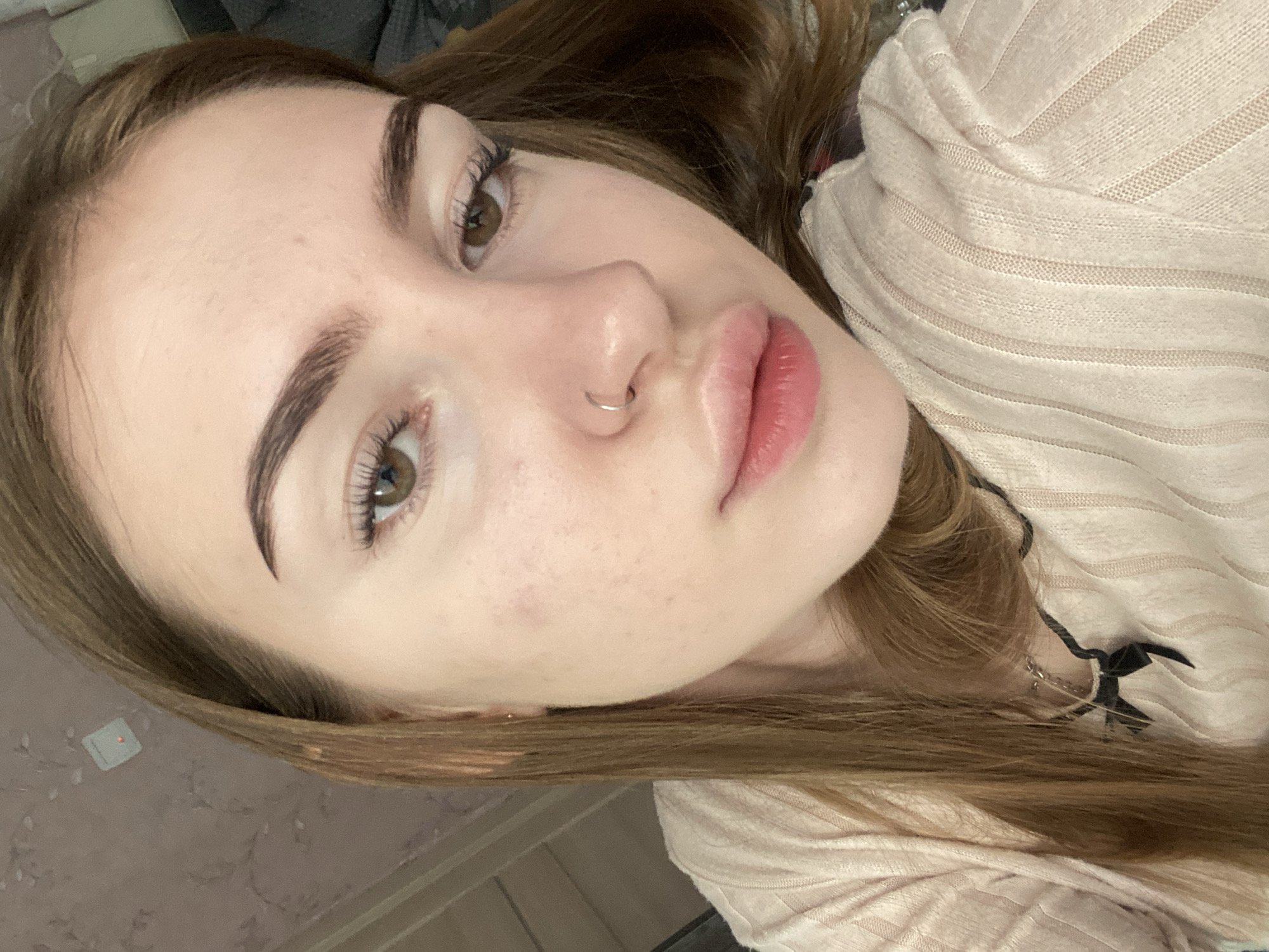 The image is a close-up, portrait-style photograph of a young woman. She has long, light brown hair that is parted and falls around her face. Her skin is fair with some visible pores and a few faint red marks on her cheek. She has well-defined eyebrows and light brown eyes with visible eyelashes. Her nose has a small, silver hoop piercing. Her lips are full and appear to be a light pink color, with one slightly more defined than the other. She is wearing a light-colored, ribbed top. A dark necklace with a black ribbon or bow pendant is visible around her neck. The background is blurred, appearing to be an indoor setting with a patterned wallpaper on the left and a darker, possibly upholstered surface on the right. There is a small, square white object with a red light attached to the wallpaper in the upper left corner of the image. The image does not provide any specific visual cues to determine its location within the city of Дьяково, Russia.