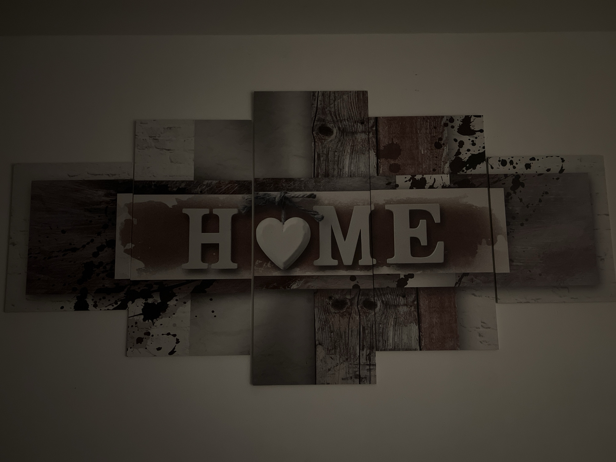 This is a digital photograph of a wall art piece displaying the word "HOME". The art is composed of multiple rectangular panels arranged in a staggered pattern. The central panel features the word "HOME" in large, white, three-dimensional block letters. The letter "O" is replaced by a white, three-dimensional heart shape, tied with a rustic rope. The background of this central panel has a reddish-brown, textured appearance with some darker splatters.
Surrounding the central panel are other panels with various textures and patterns. Some panels depict distressed wood grain, others have a light gray or white distressed brick pattern, and some feature dark splatters on a lighter background, creating a modern, abstract aesthetic. The art is mounted on a plain, light-colored wall, which appears to be the interior wall of a residence. There is no indication of the specific location in Bad Zwischenahn, Germany, nor are there any people or actions depicted. The lighting is dim, casting shadows and highlighting the textures of the artwork.