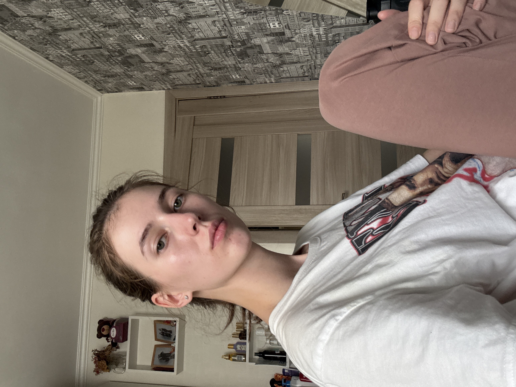 The image depicts a young woman with light brown hair pulled back, wearing a white t-shirt and pink loungewear. She is seated indoors, with her legs crossed. The background shows a wall with a black and white city-themed wallpaper, a wooden door, and a shelf with decorative items including framed pictures and a teddy bear. Based on the decorative elements and the style of the interior, it is plausible this scene is located in Perm, Russia.