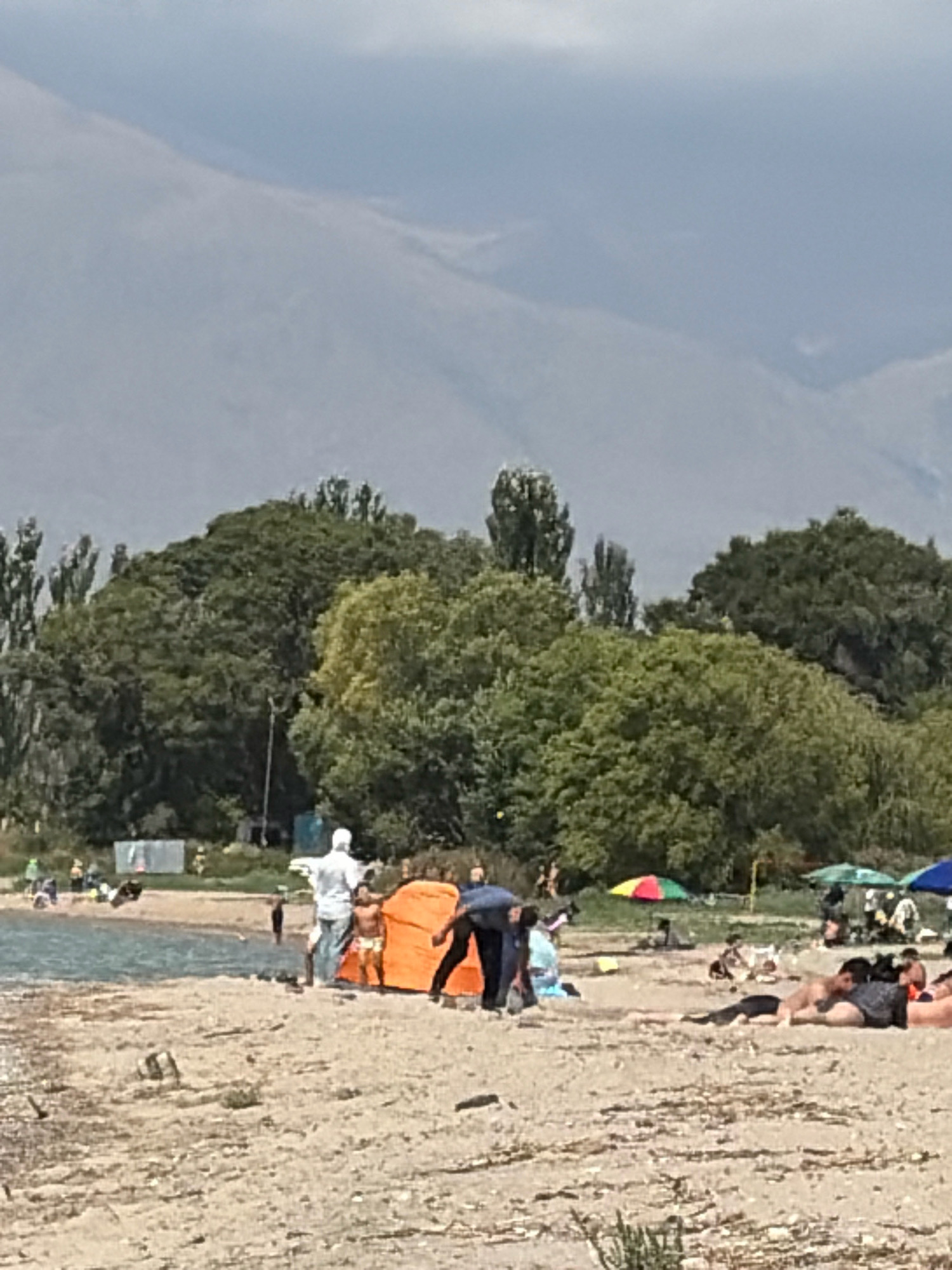 The image shows a beach scene with people relaxing and playing near the water. In the foreground, sand is visible with some scattered debris. In the midground, several people are on the beach. One person in a white shirt stands near an orange tent, facing away from the camera, while a small child in shorts stands in front of the tent. To the right, a person in dark clothing is bending over. Further back, other individuals are lying on the sand, some under beach umbrellas. The water's edge is visible, with ripples indicating gentle waves. In the background, there are trees and hills under a cloudy sky. A few beach umbrellas are also scattered across the beach.