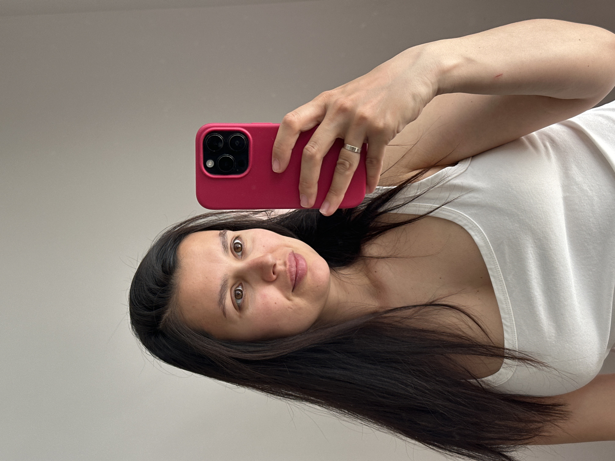 A woman is taking a selfie. She is holding a pink phone case with a black phone inside. She has long, dark hair and is wearing a white tank top. She is wearing a silver ring on her left hand. The background is a light grey wall. It is not possible to determine the location in the city of Chaikovsky, Russia from the given image.